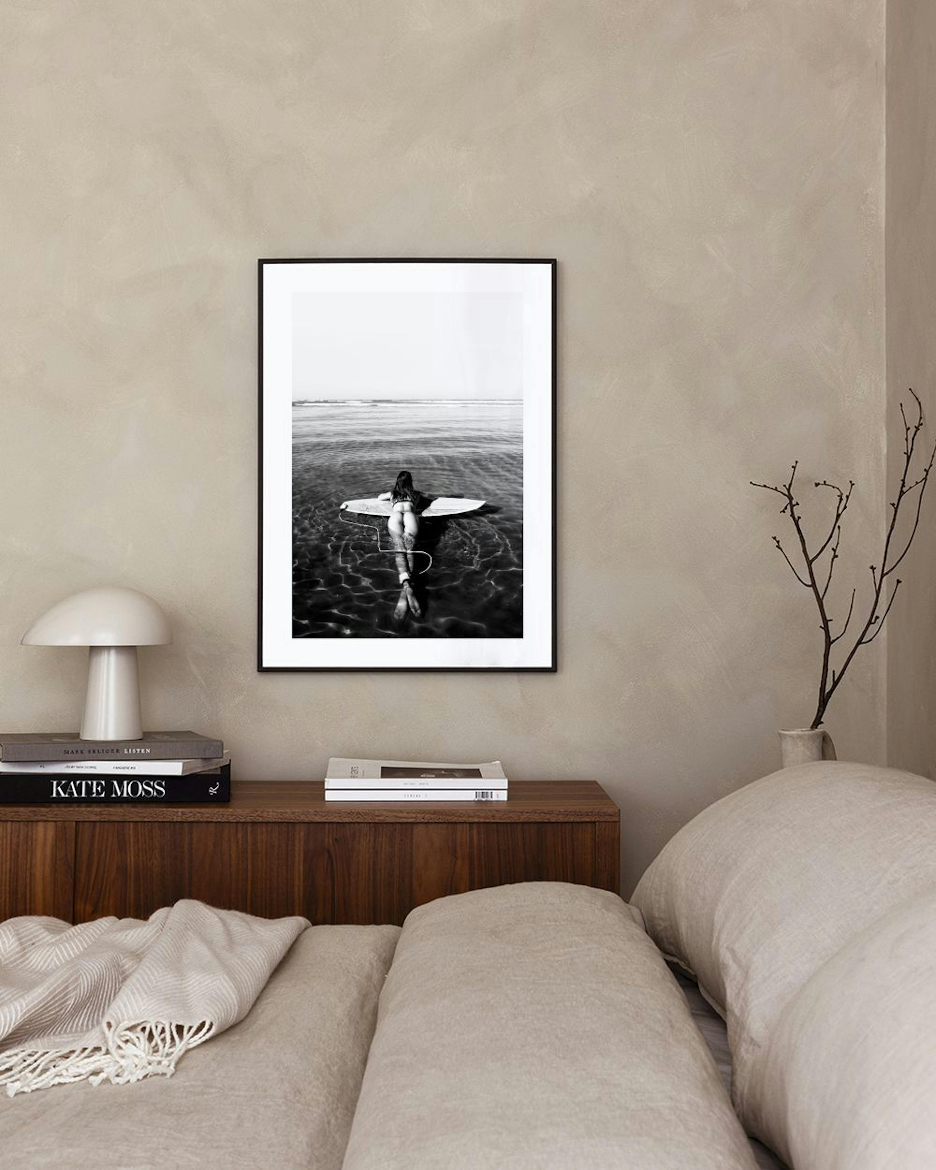 Black and white print of a person on a surfboard in water, hanging above a wooden desk with books and a vase.