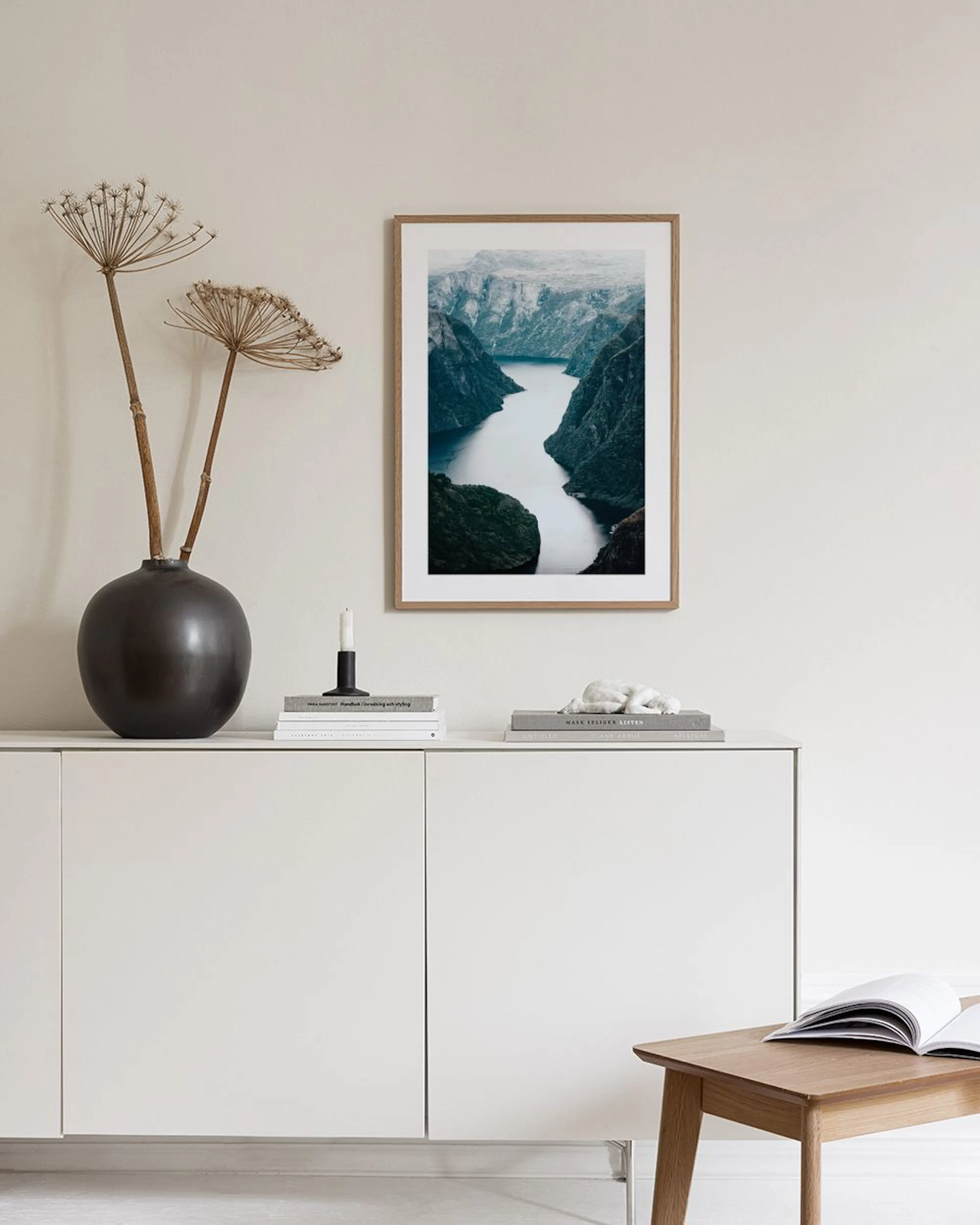 Poster con fiordo norvegese, pareti rocciose e acqua grigia, appeso in un moderno soggiorno con pouf e tavolino.