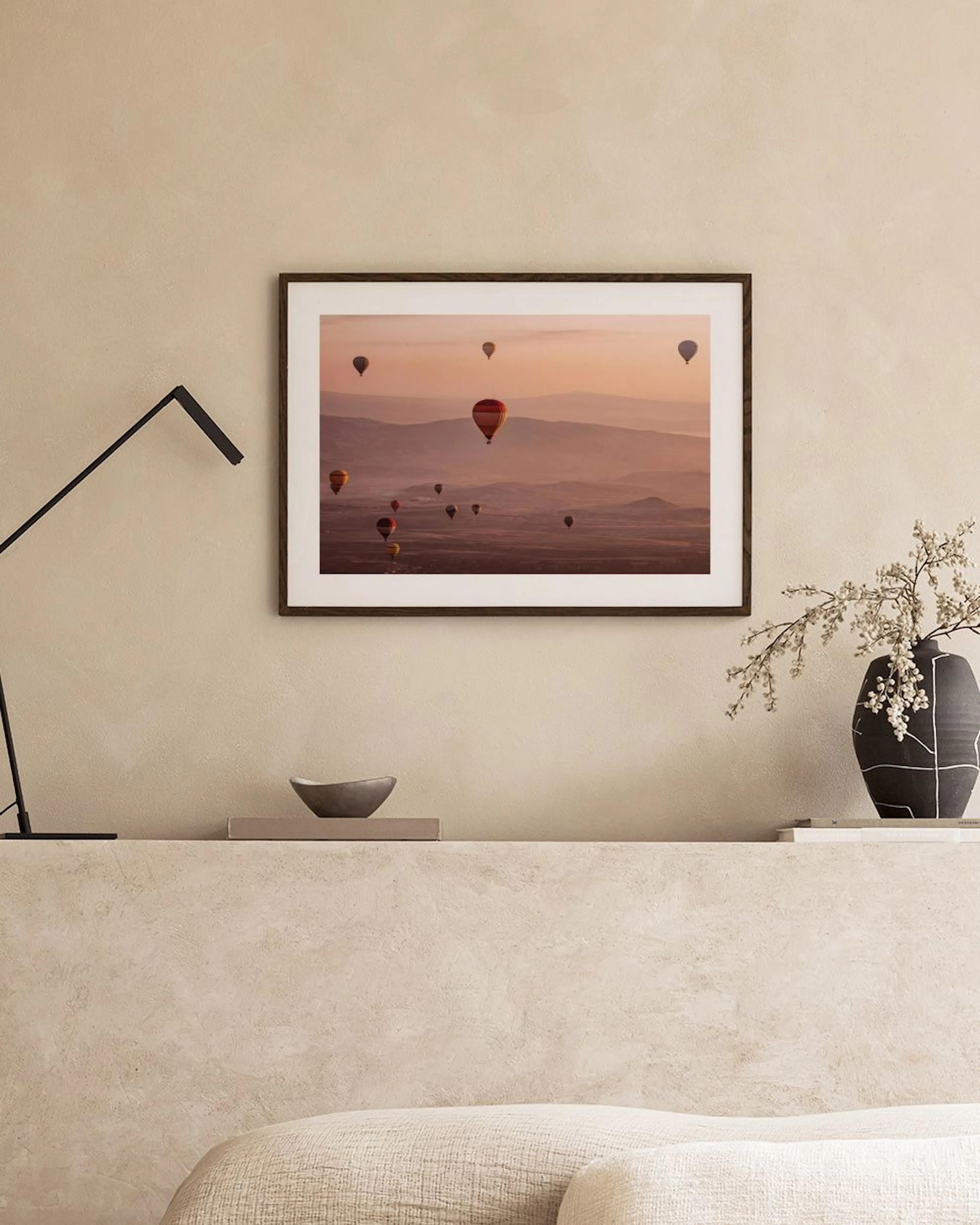Poster con mongolfiere su paesaggio montuoso, appoggiato su una panca con vaso e libri in una stanza moderna.