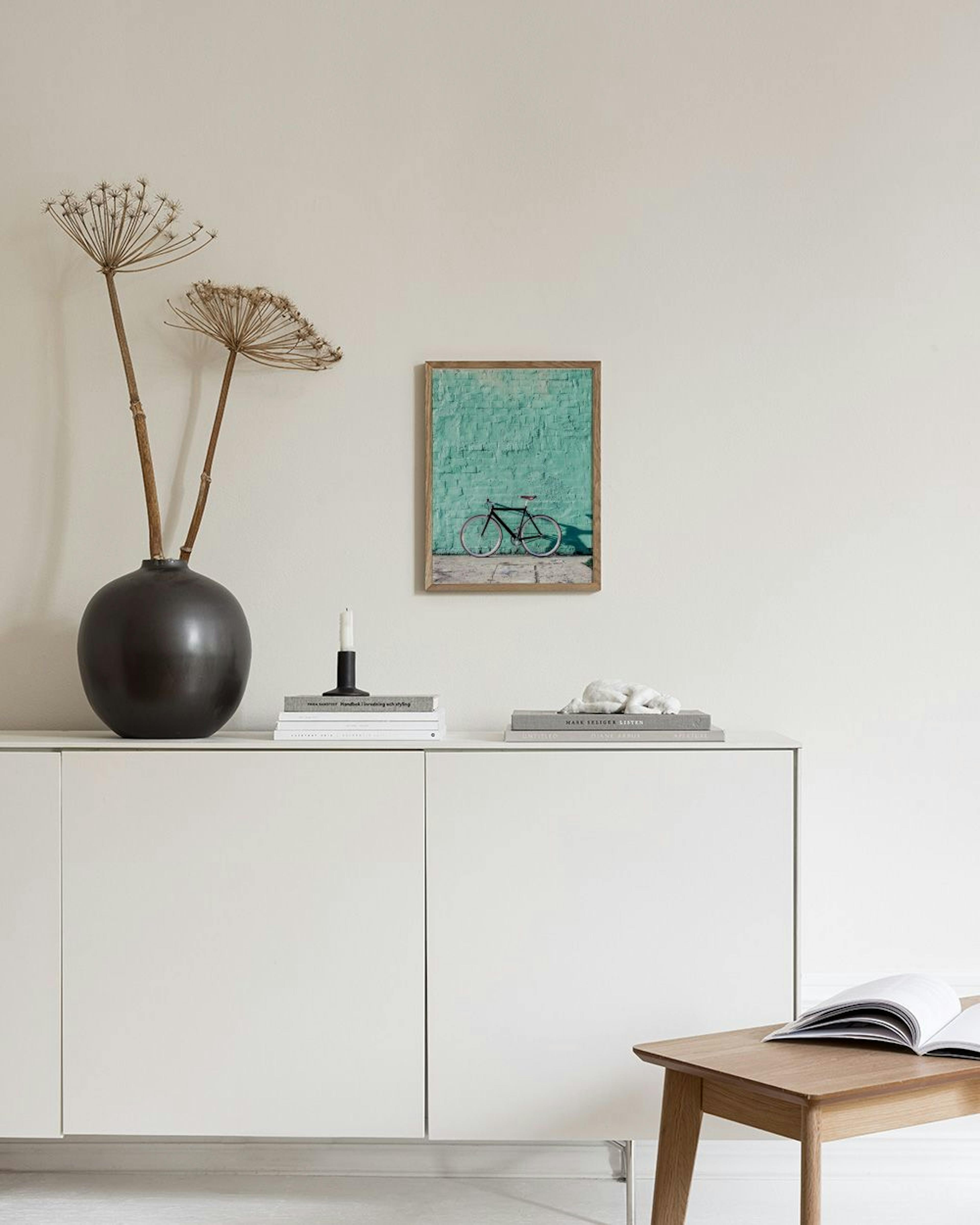 A gallery wall of six teal and pink prints hangs above a velvet ottoman with pillows, beside a bright window.
