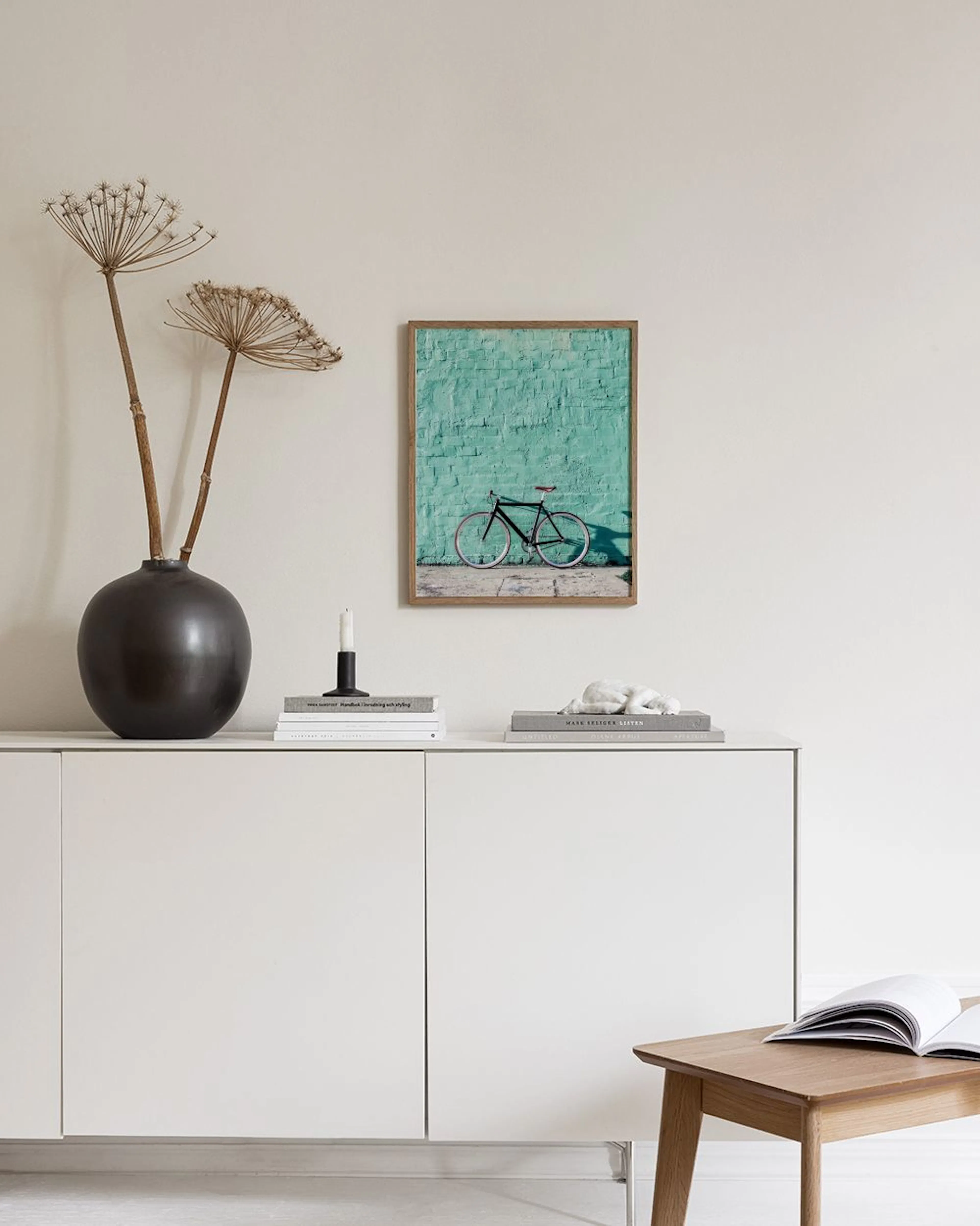 A gallery wall of six teal and pink prints hangs above a velvet ottoman with pillows, beside a bright window.