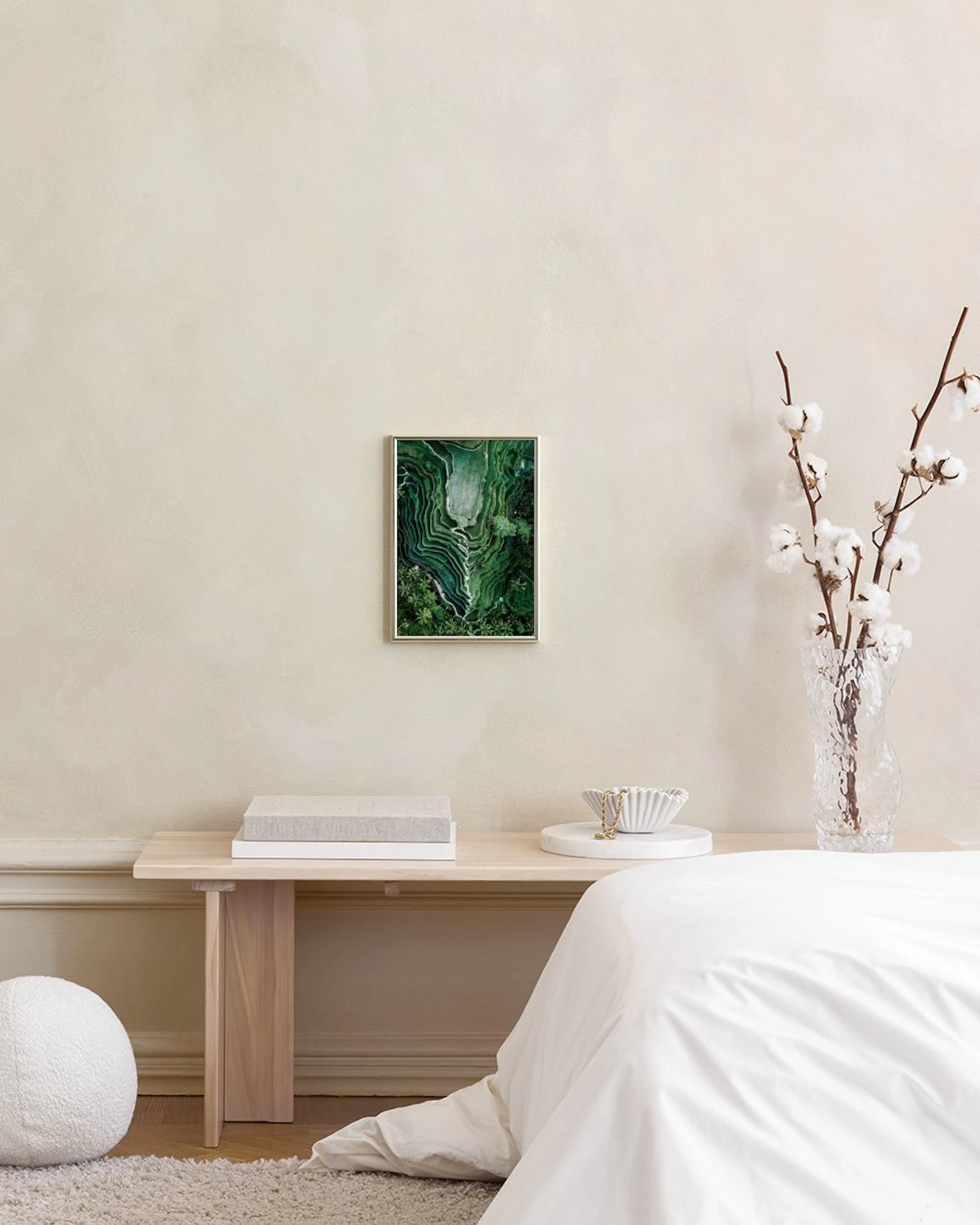Plakat med flyfoto av grønne, terrasserte rismarker og palmer, henger på en hvit vegg over et skap med vase.