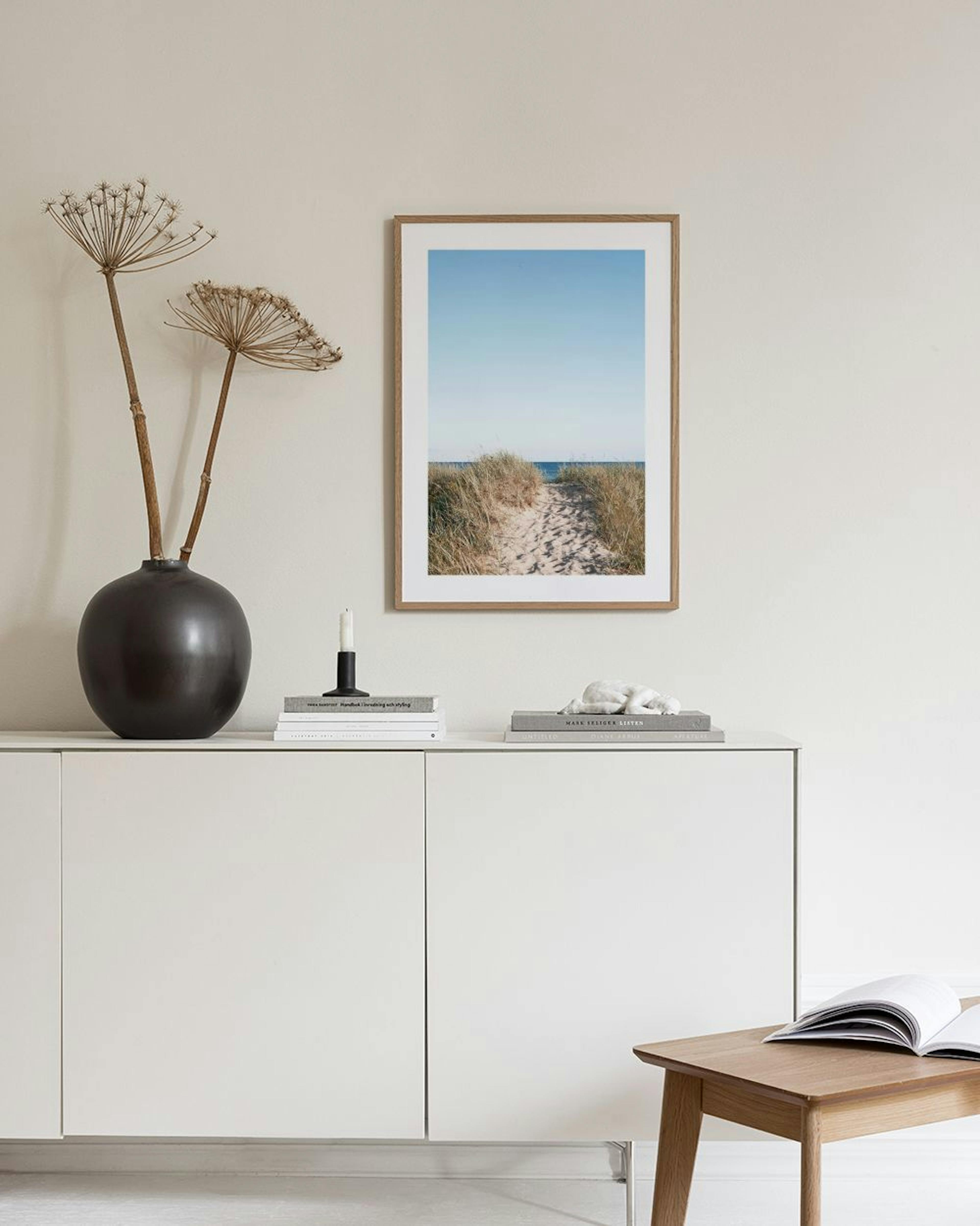 Affiche minimaliste dun chemin sablonneux vers la mer, encadrée et exposée sur un meuble blanc avec un vase et des livres.