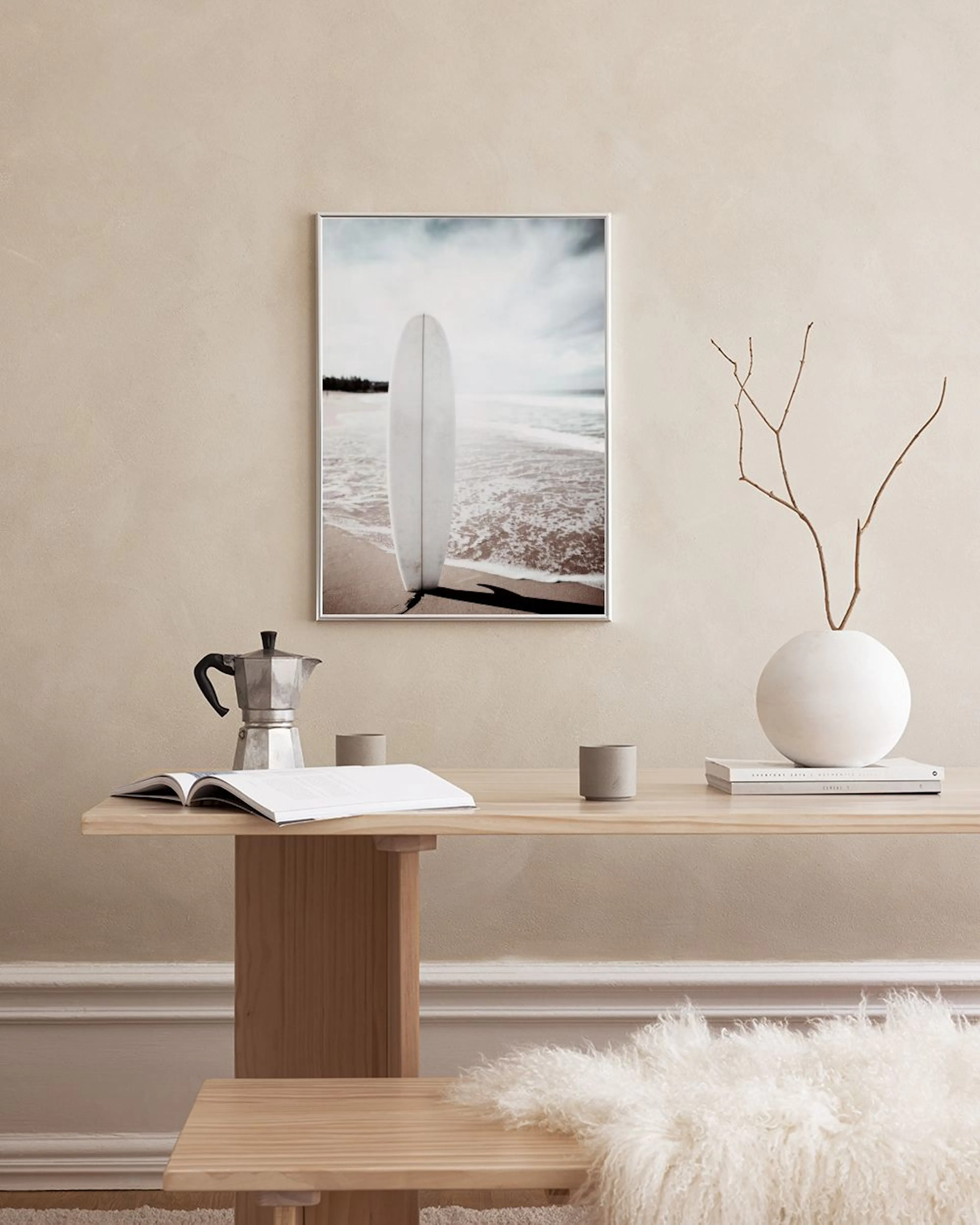 A print of a white surfboard on a sandy beach with ocean waves, hanging above a pink velvet sofa with pillows.