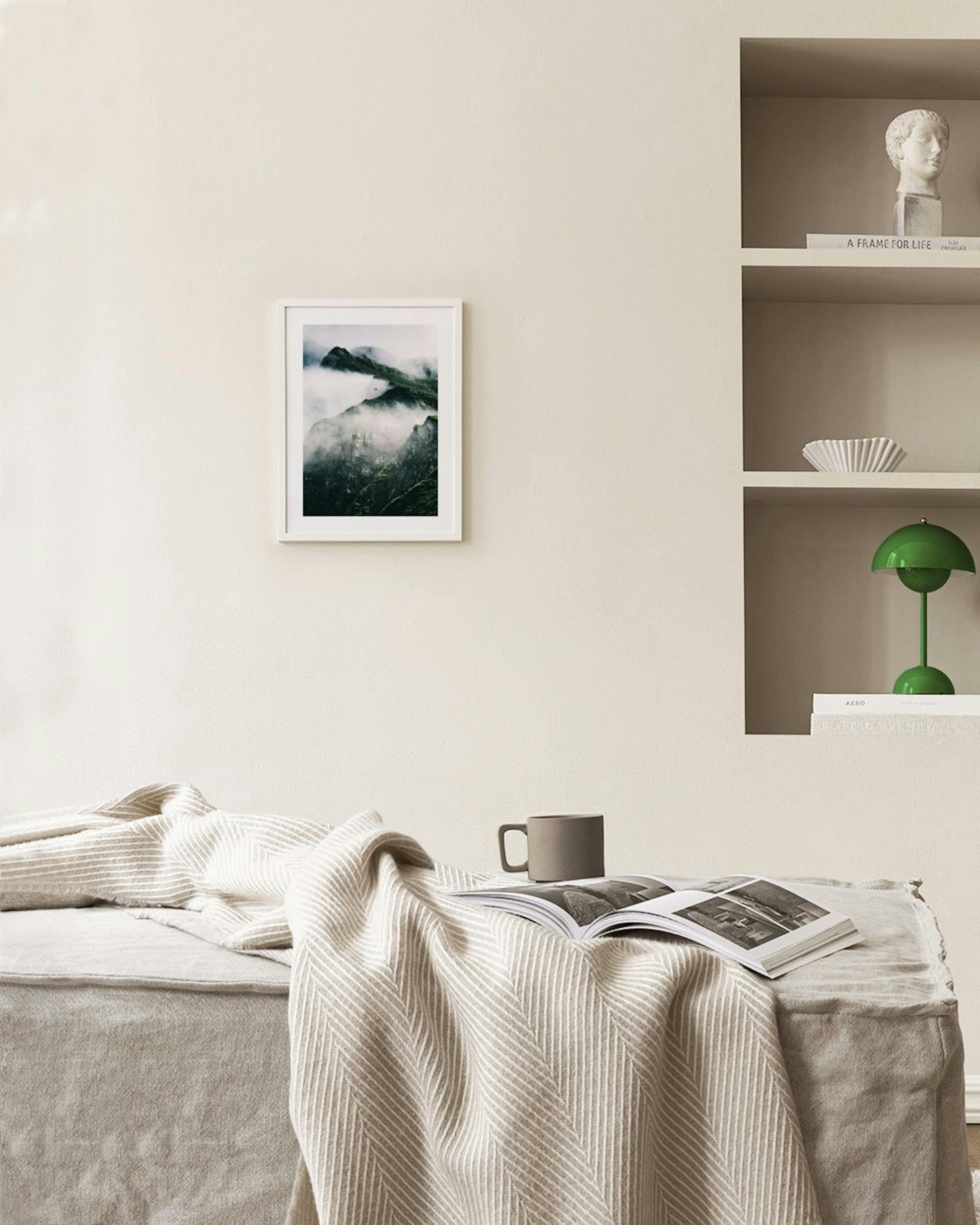 A print of dark green mountains partially covered by white clouds, hanging above a white console table with a brown chair.