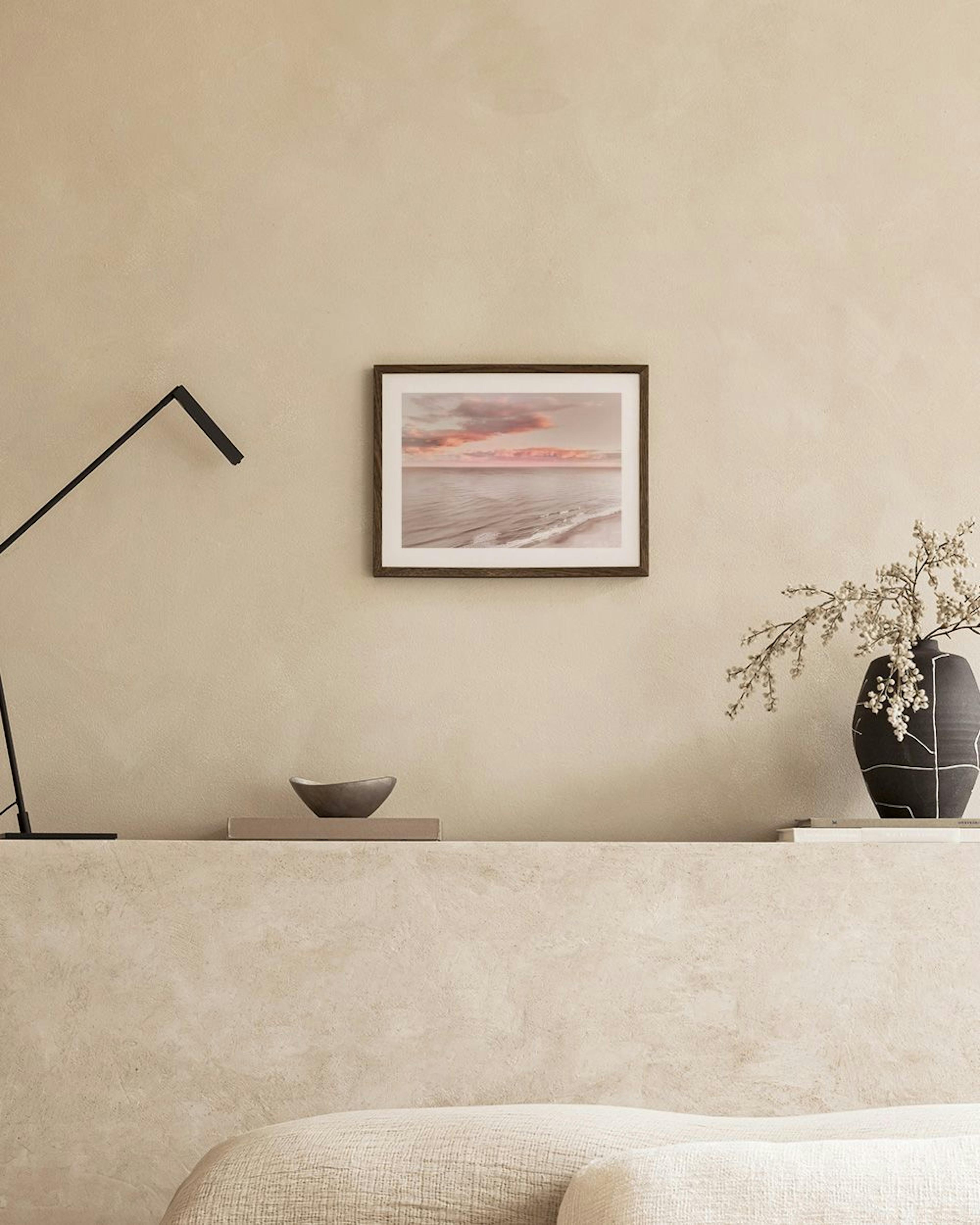 A print of a tranquil beach sunset with pink and grey clouds over the ocean, hanging in a minimalist living room.