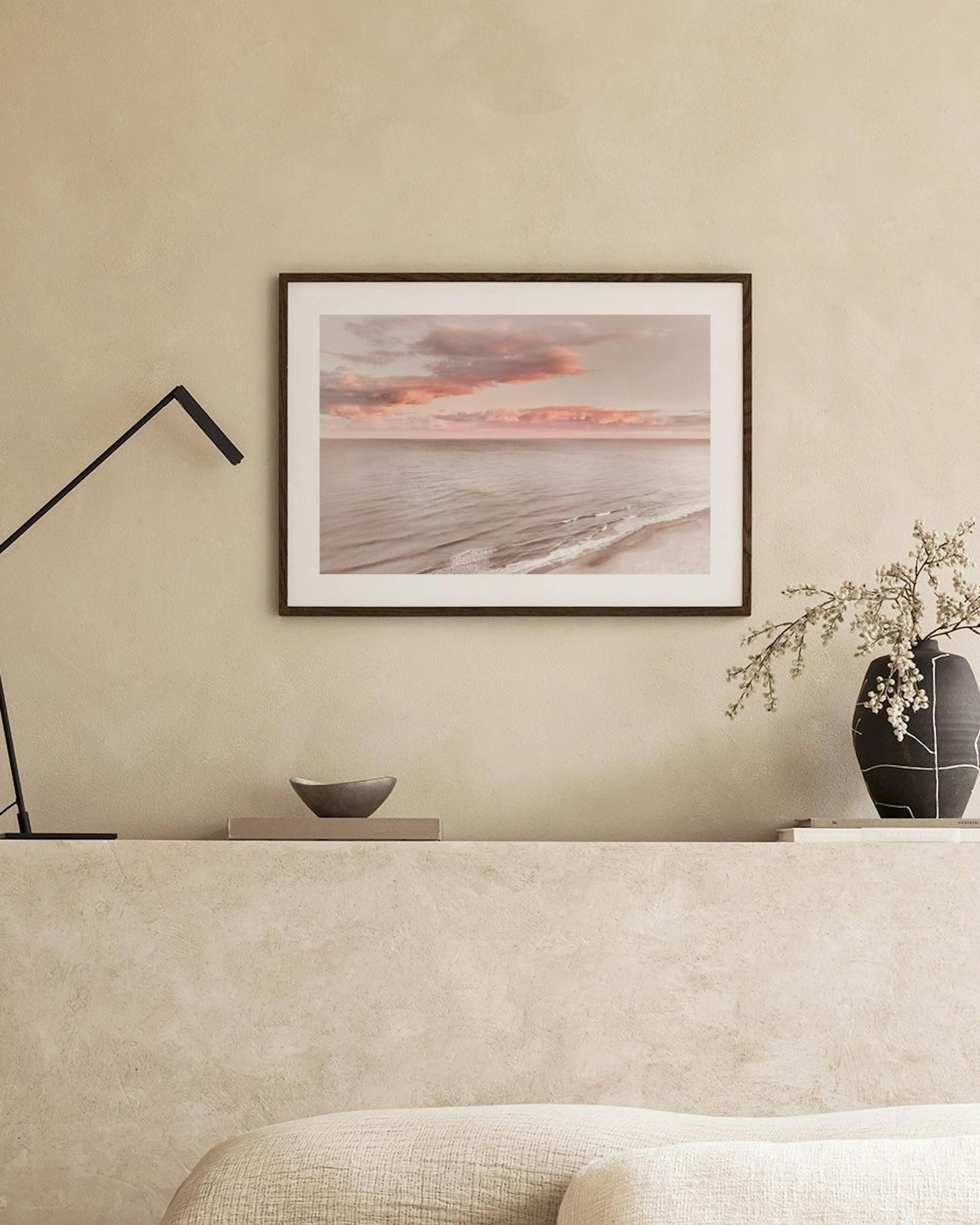 Print of a calm ocean with pink and beige clouds at sunset, displayed above a grey coffee table in a living room.