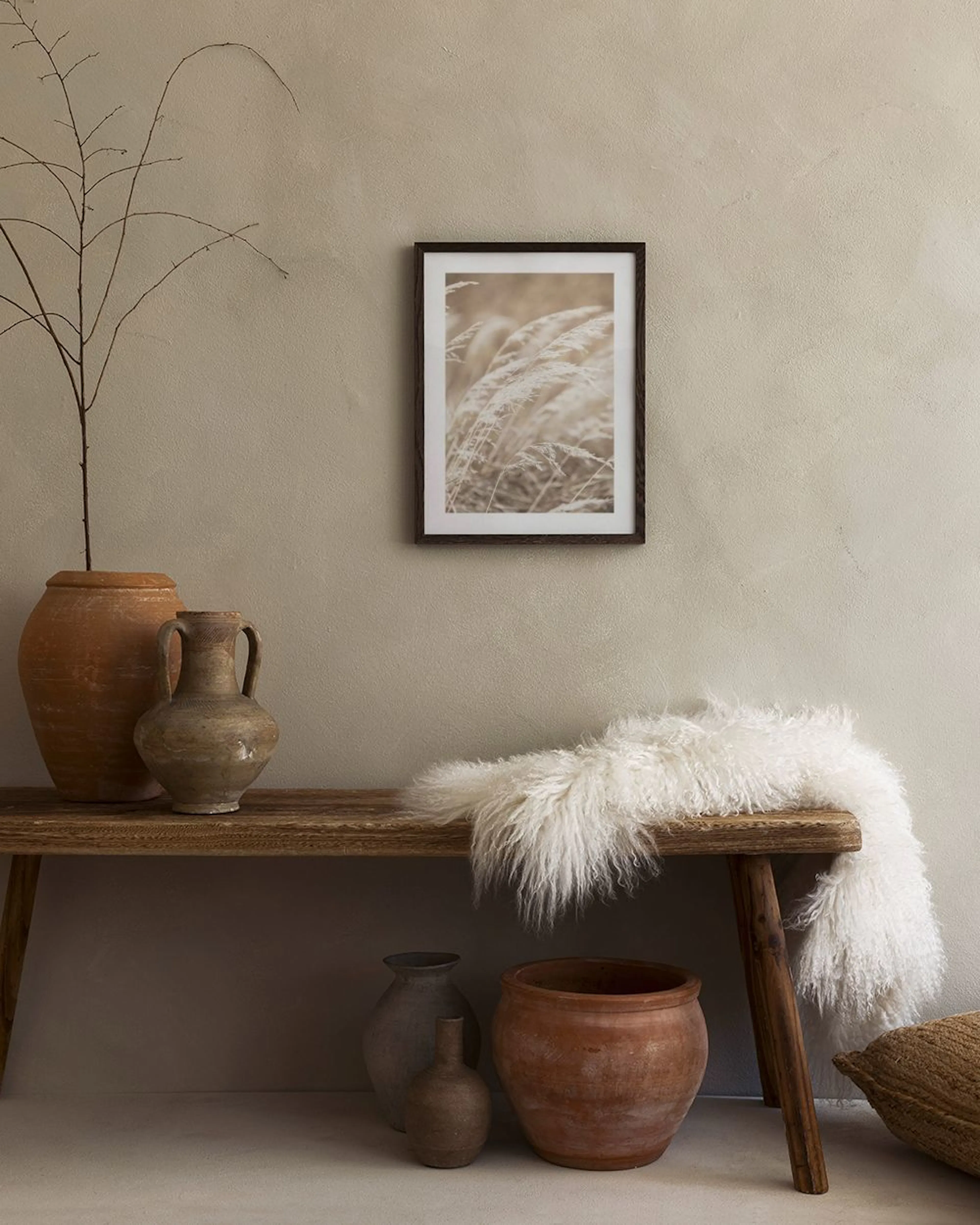 Poster mit hellem, windbewegtem Gras, hängt über einem braunen Pouf neben einem Beistelltisch mit Lampe.