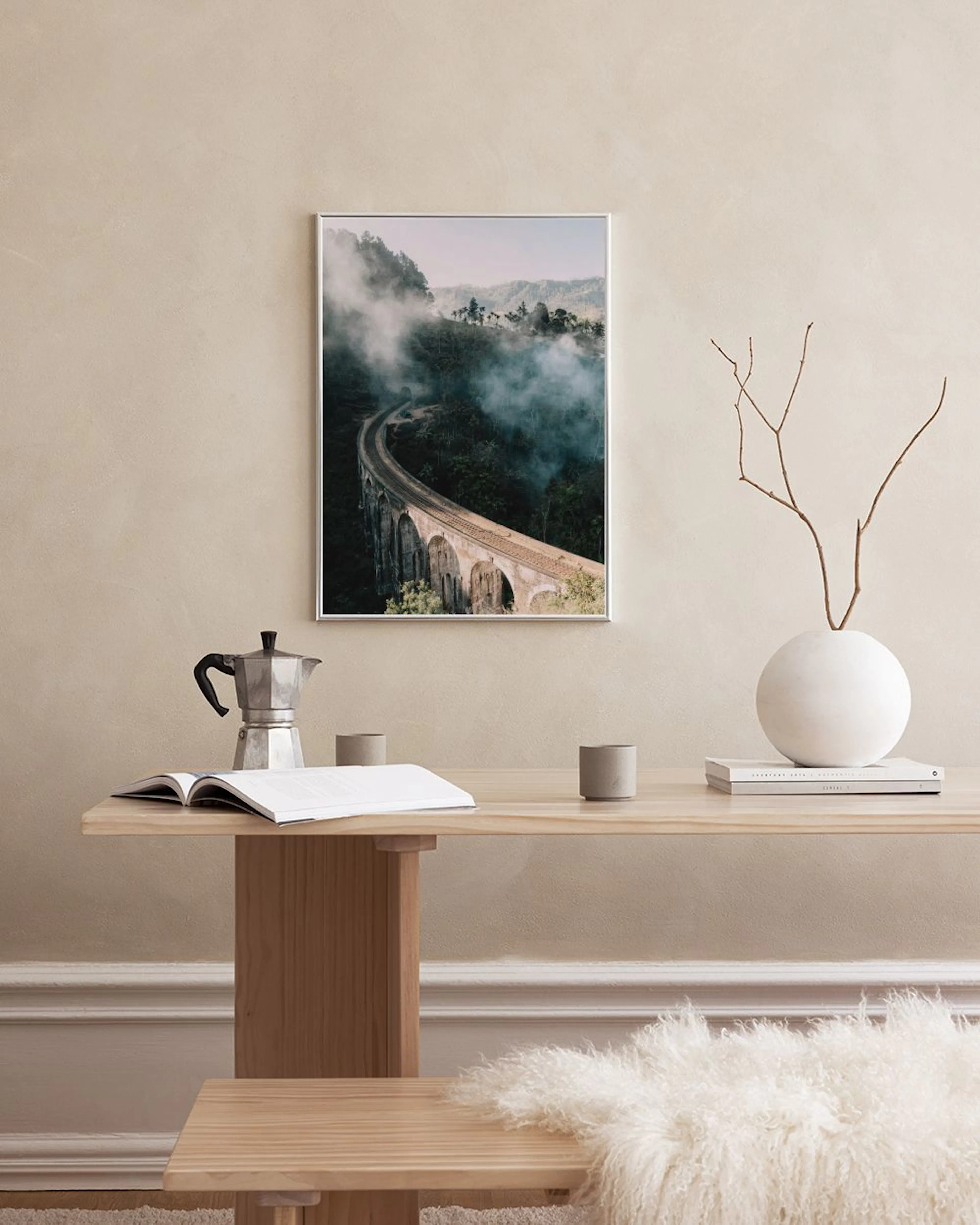 Poster av tegelbro i dimmigt berglandskap, hängande ovanför en träbänk med växter och inredning.