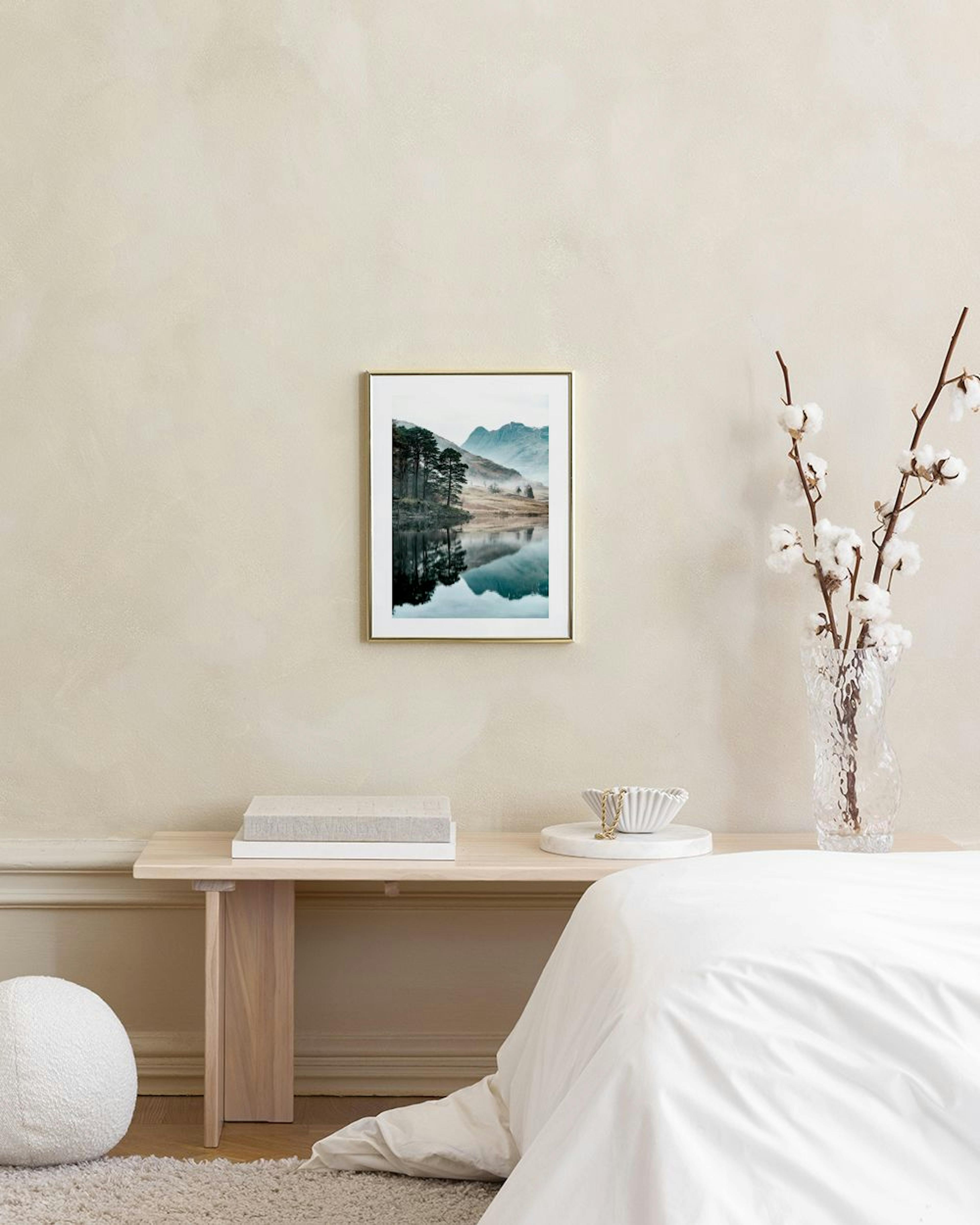 Póster de un paisaje brumoso con lago y árboles, exhibido sobre un mueble blanco con un jarrón y objetos decorativos.