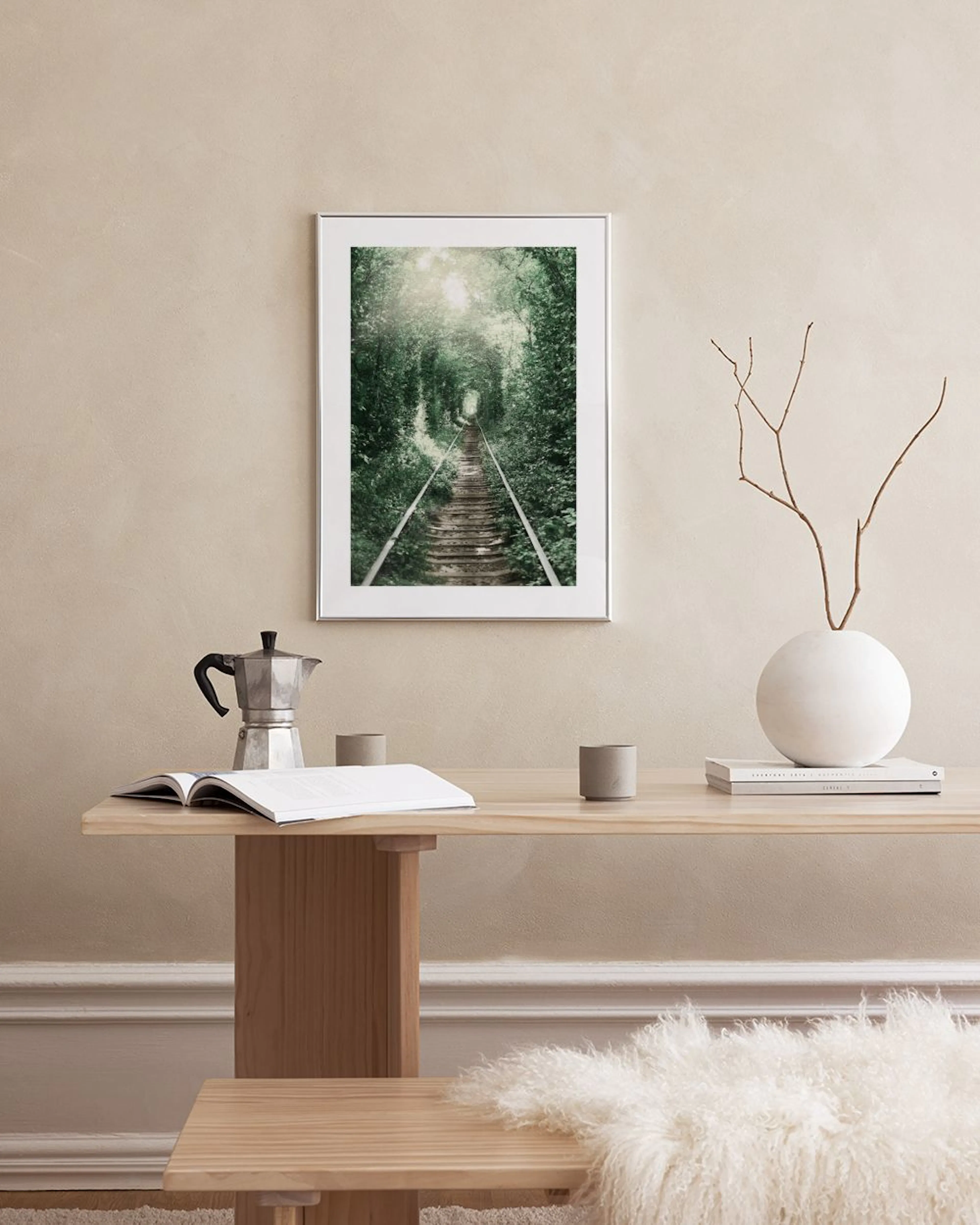 Plakat med jernbanespor gennem grøn løvtunnel, hængende over et lyst skrivebord med kaktus og magasin.
