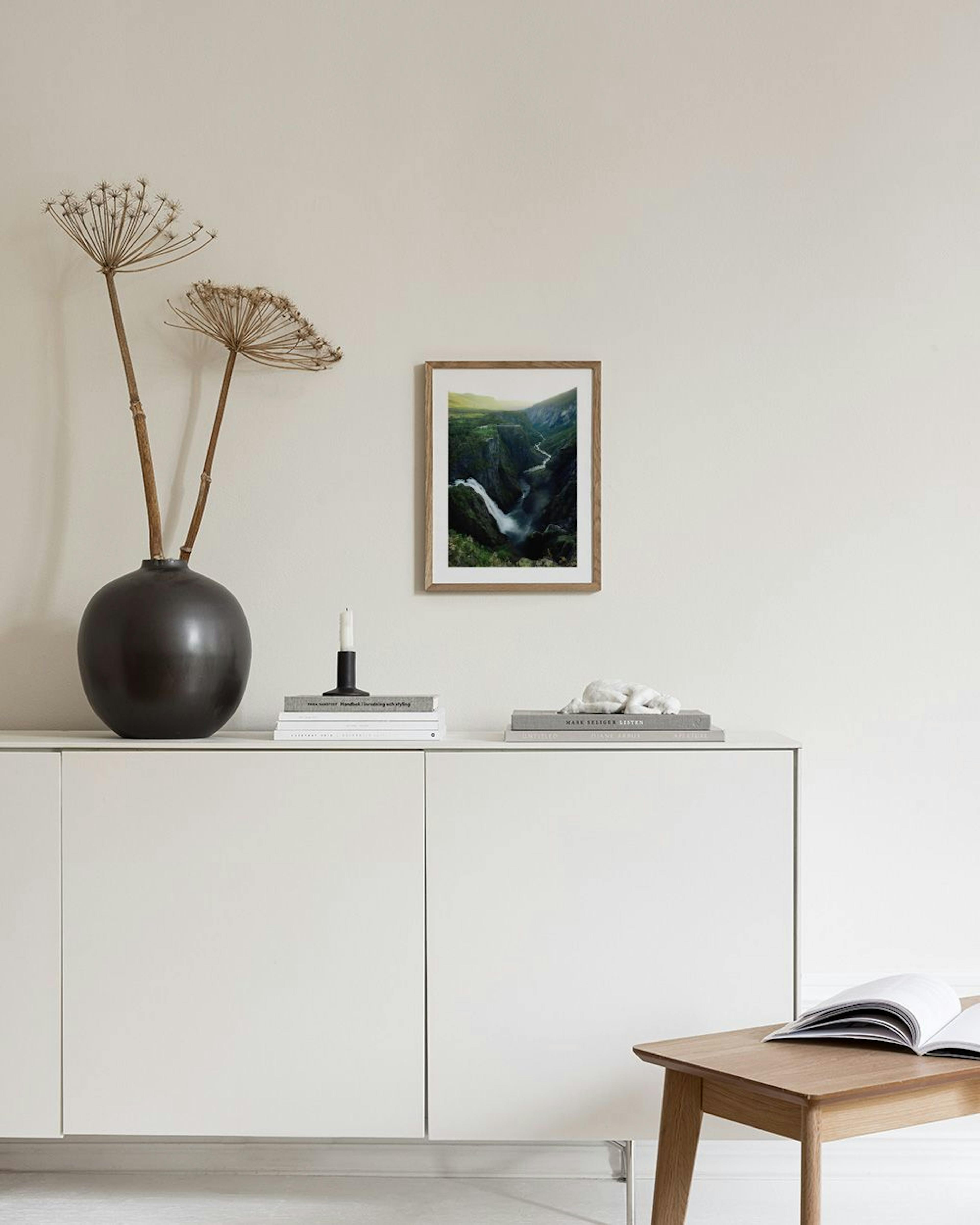 A print of a dramatic waterfall in a lush green canyon, hanging in a bright room above a white cabinet with a grey vase.