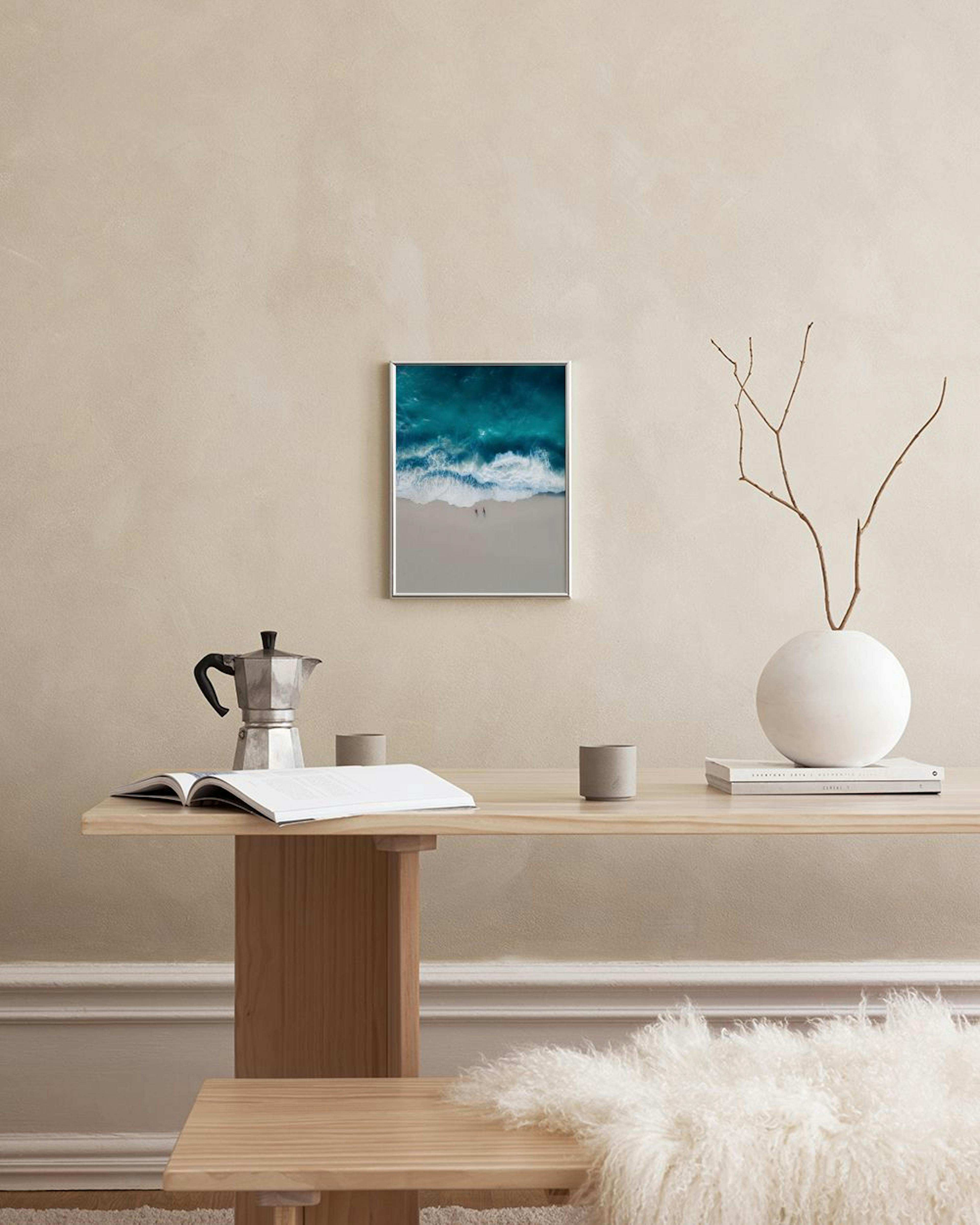 Poster eines türkisen Meeres mit Wellen und zwei Personen am Strand, über einem weißen Schrank mit Vase und Stuhl.