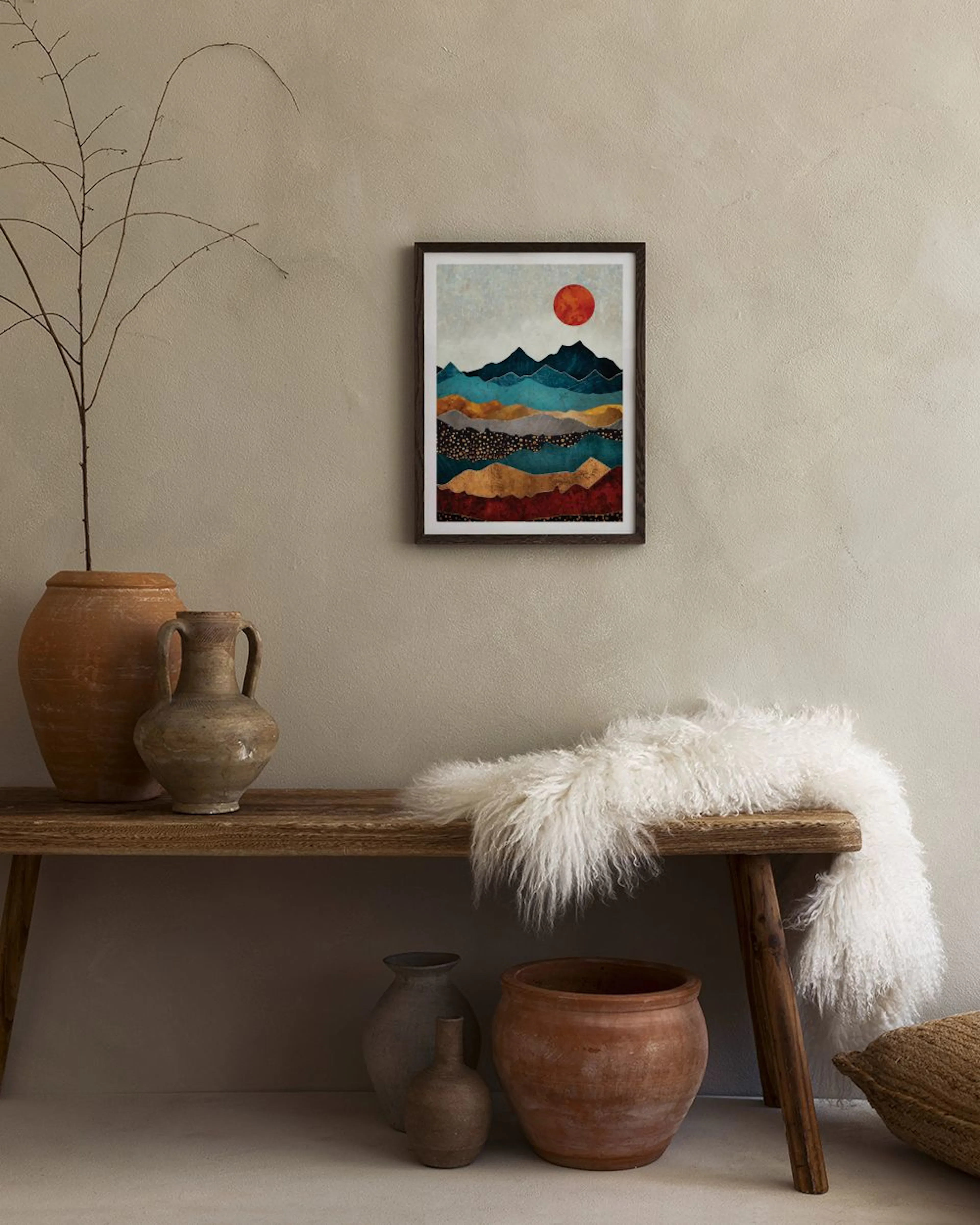 A print of abstract mountains in blue, grey, gold, and red hues with a red sun, hanging above a brown ottoman.