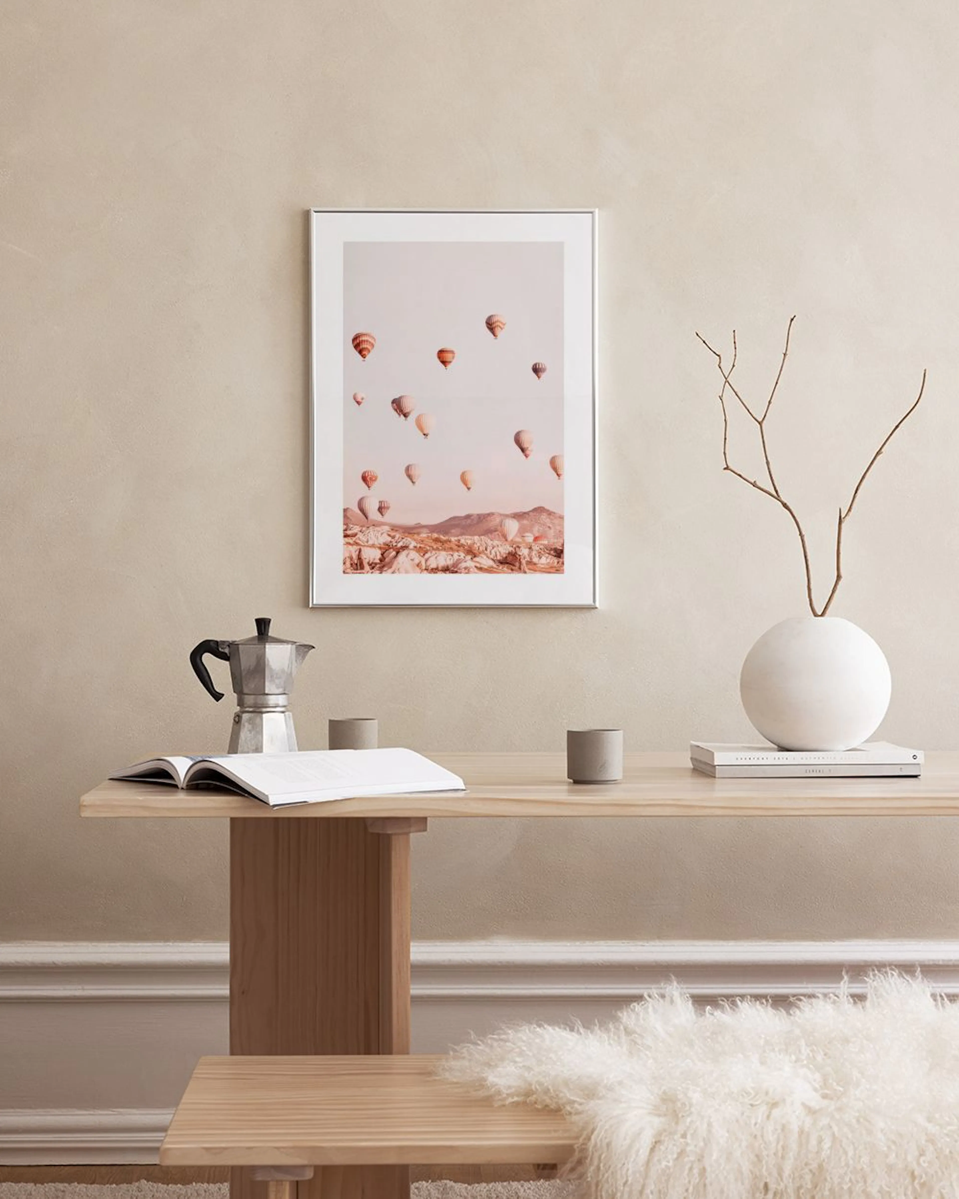 Poster mit Heißluftballons über einer Landschaft in sanften Erdtönen, hängt über einem Pouf in einem modernen Wohnzimmer.
