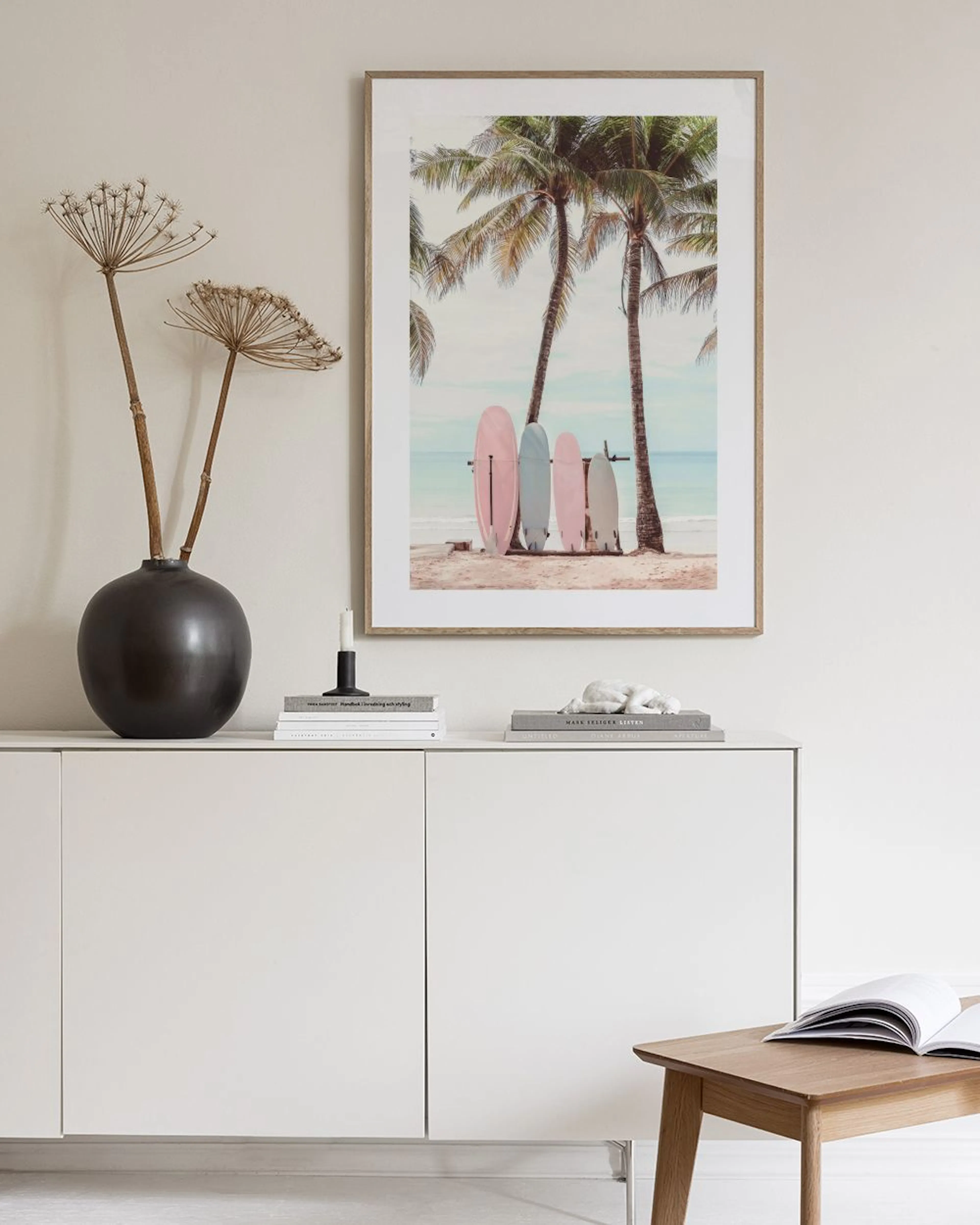 Poster mit rosa und hellblauen Surfbrettern unter Palmen am Strand, über einem weißen Sideboard mit Vase und Buch.