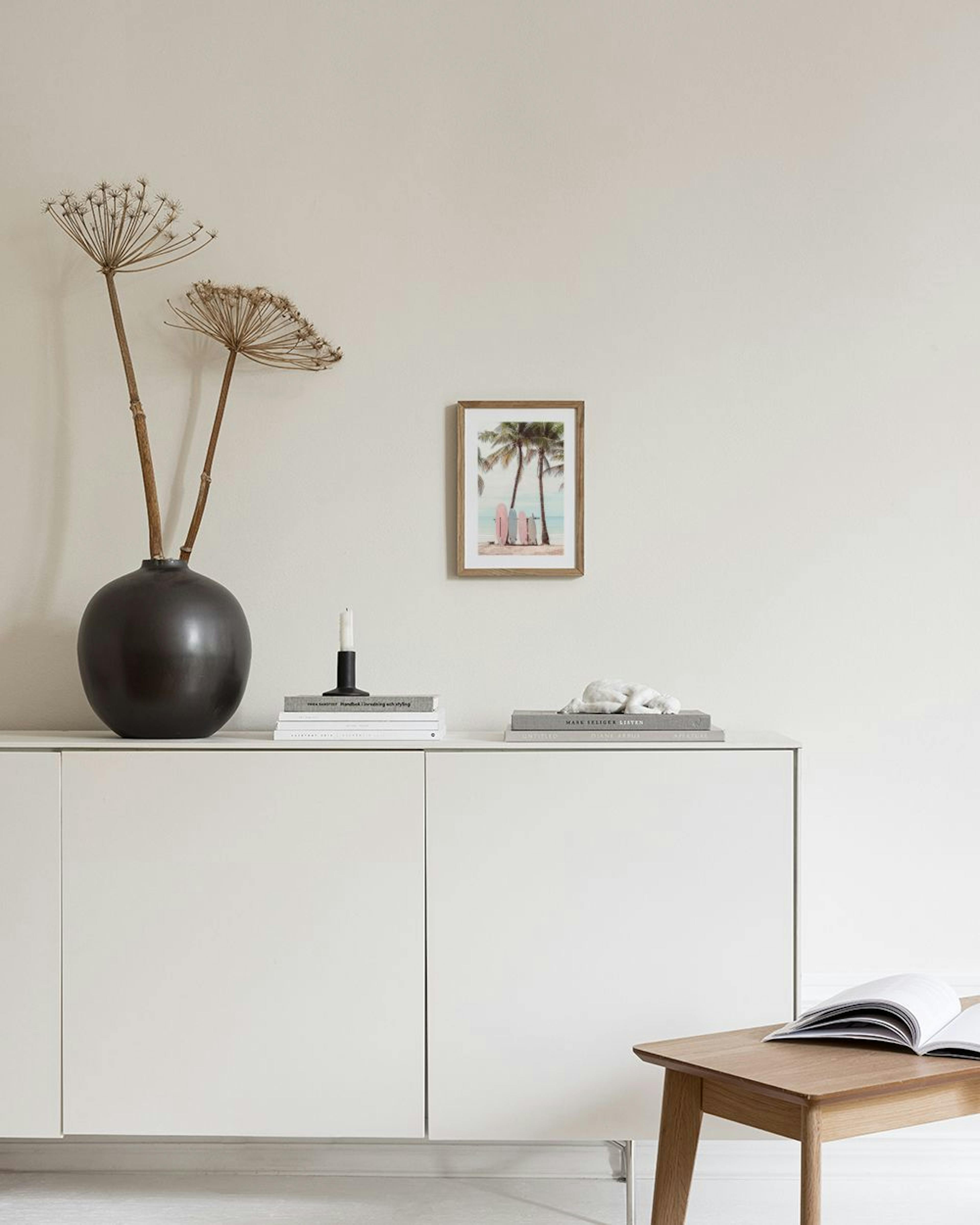 A print of pink and blue surfboards on a sandy beach with palm trees, displayed above a white console table with a vase and book