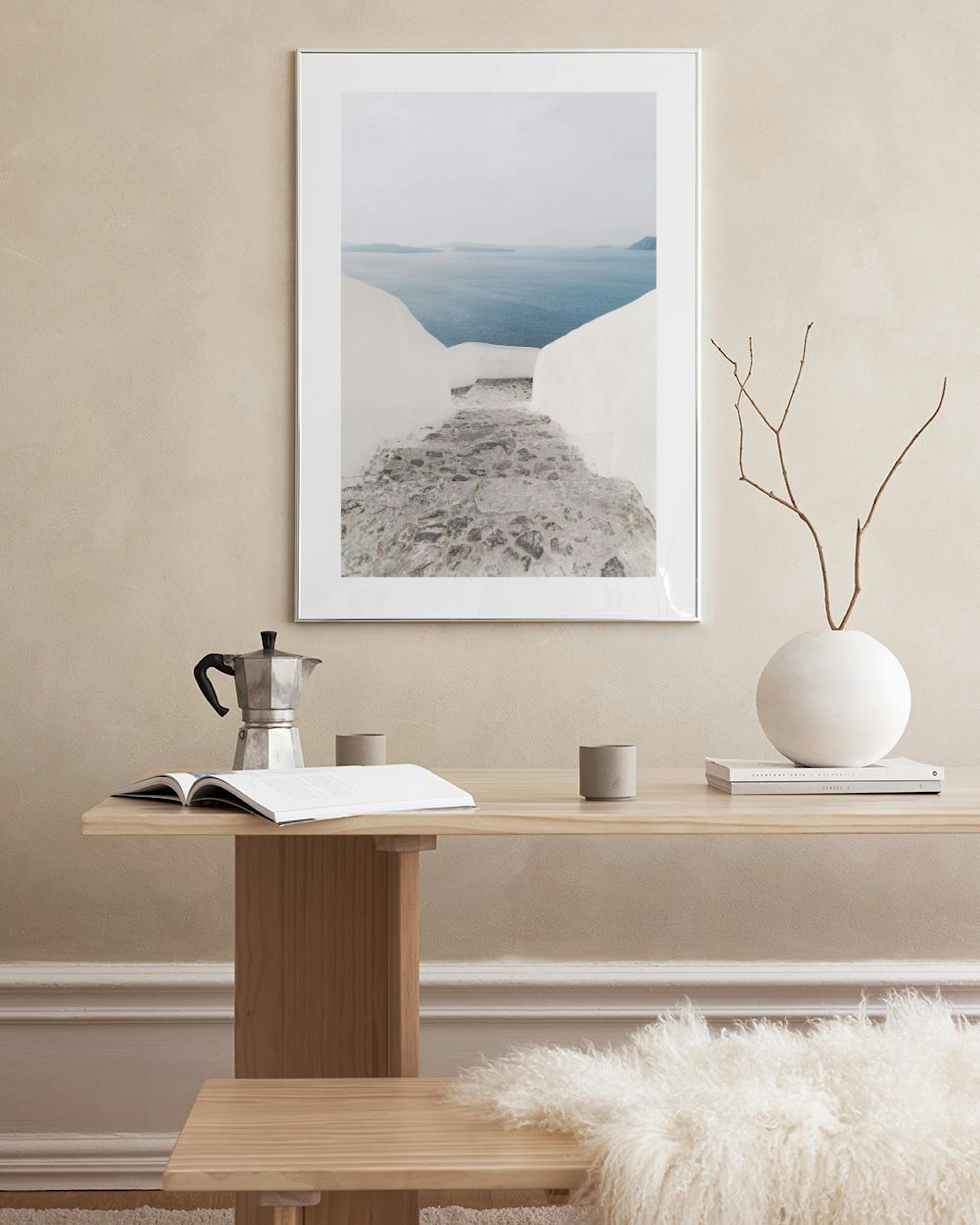 Print of a white stone path leading to a blue ocean view, hanging above a rustic wooden bench with decorative vases and baskets 