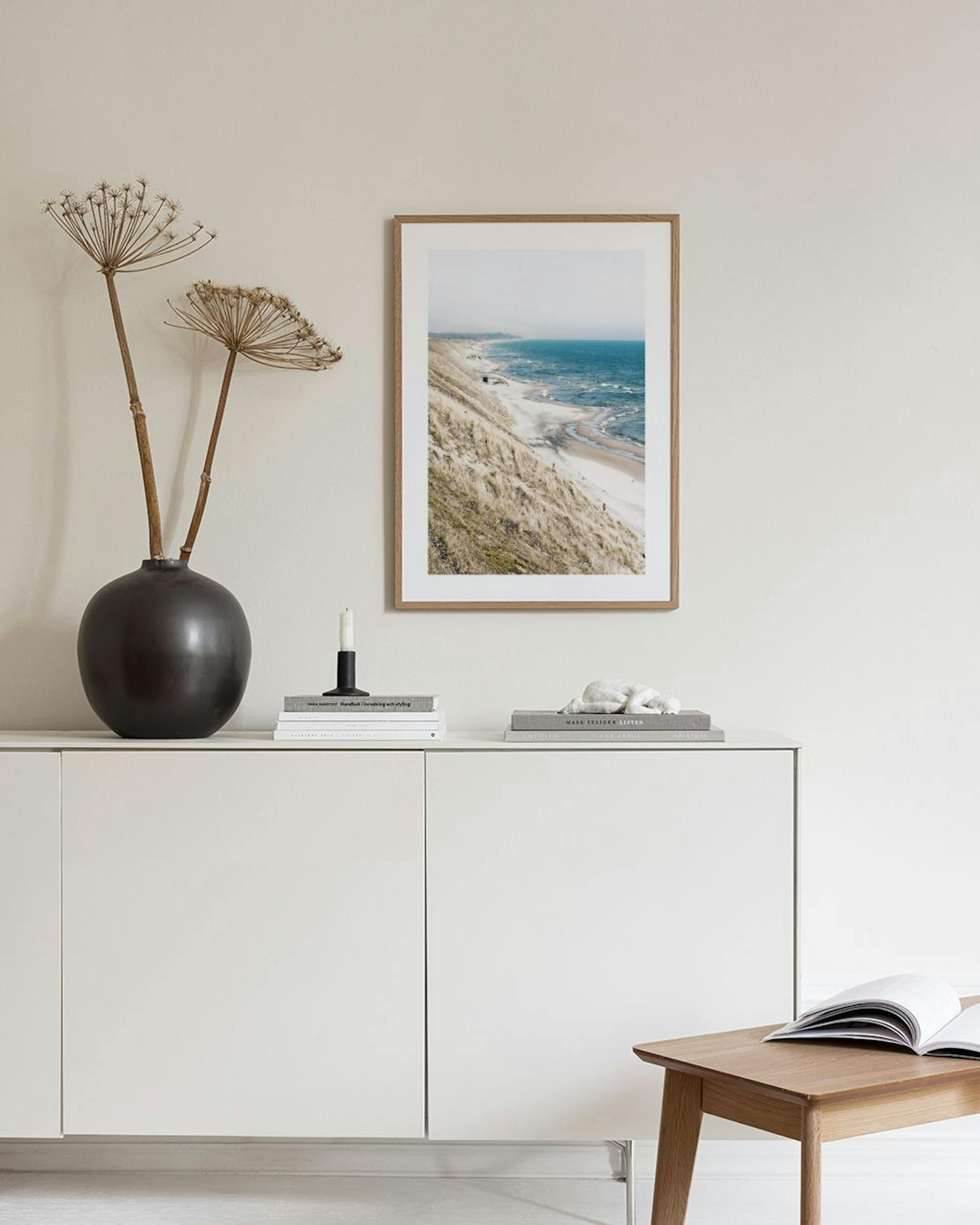 Poster mit Küstenlandschaft, Sanddünen und blauem Meer, hängt über einem Hocker mit Büchern in einem schlichten Raum.