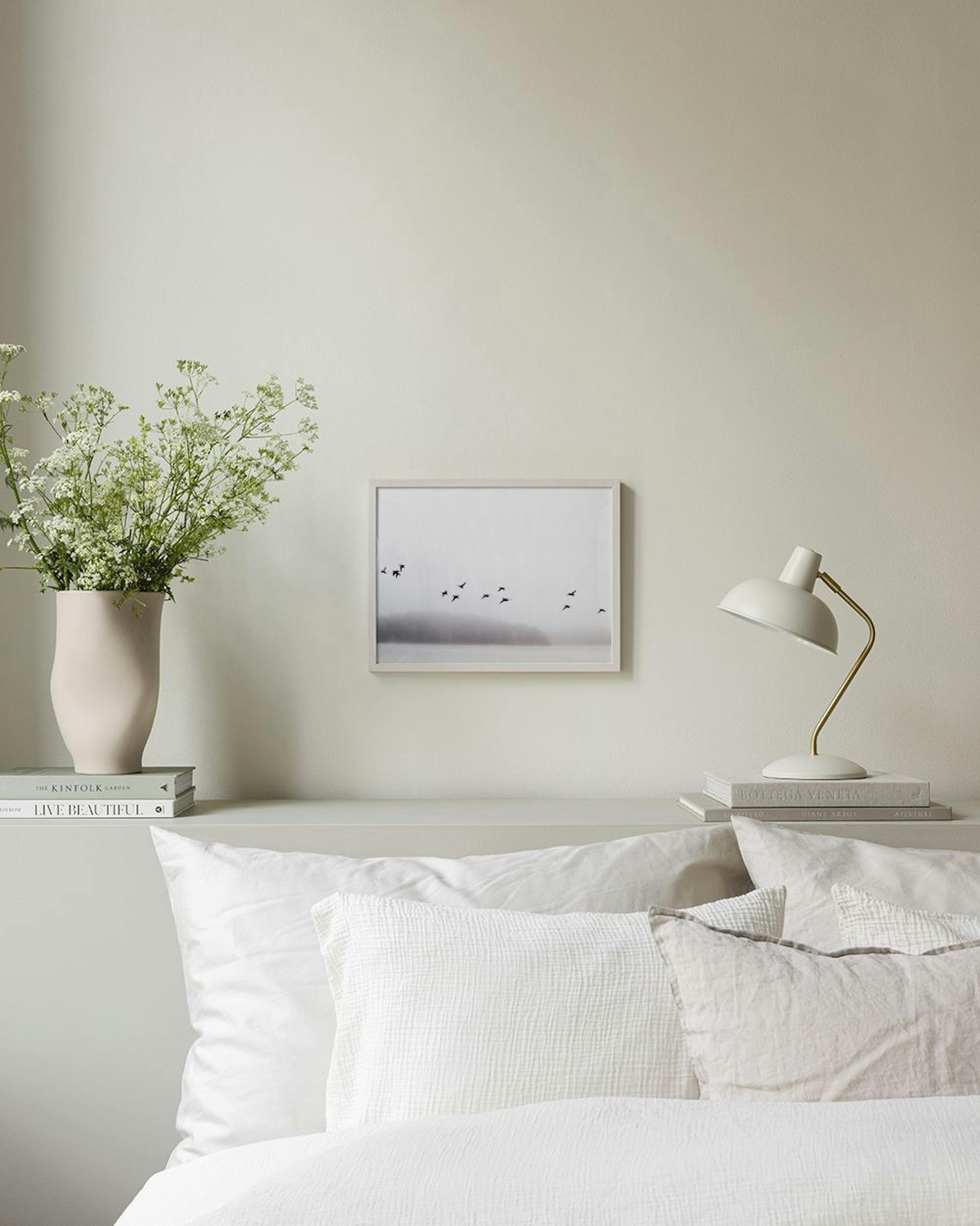 Poster med fåglar över dimmigt landskap och vatten, hängande ovanför en gråblå skänk med böcker och vaser.