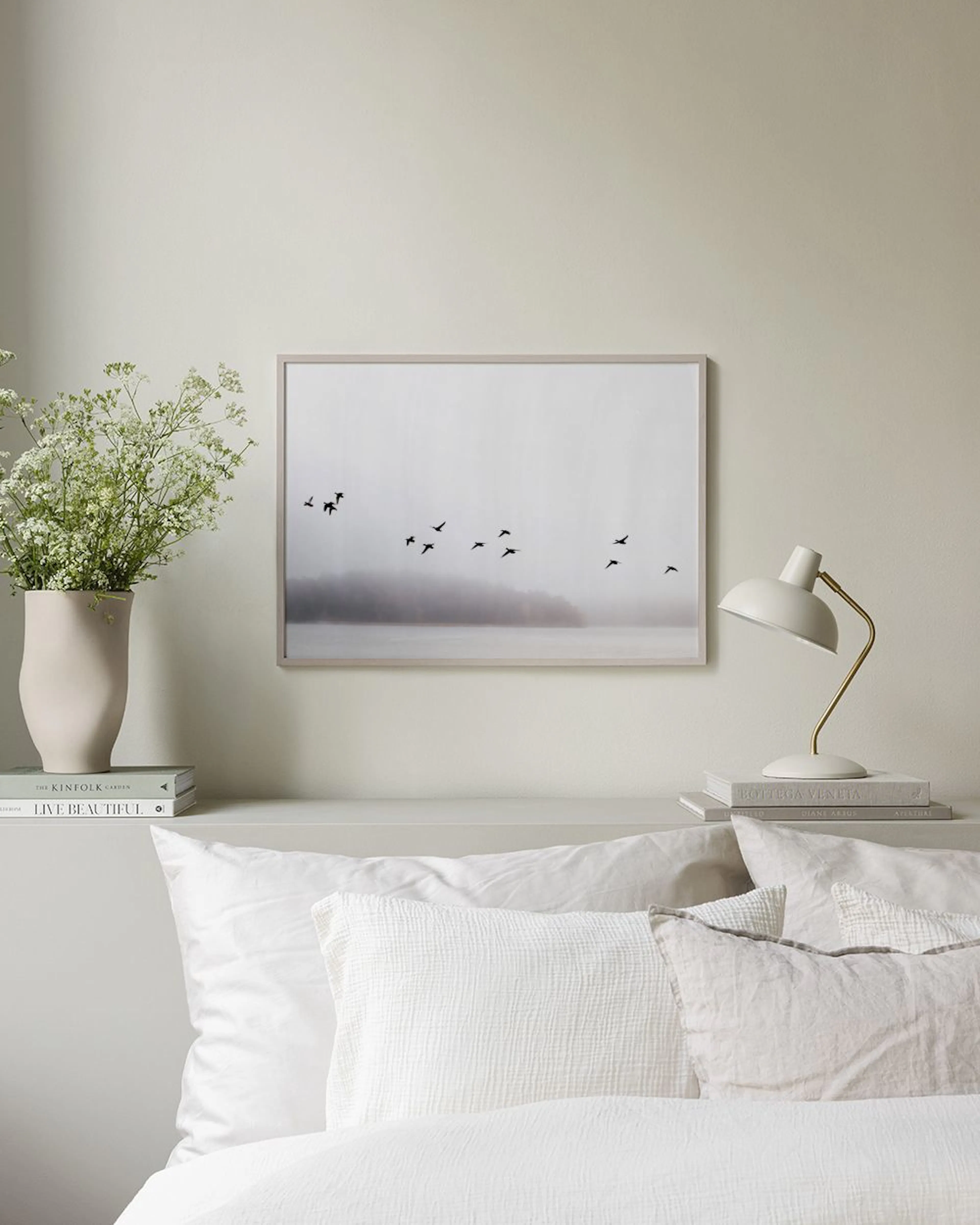 A print of black birds flying over a misty lake with trees, displayed above a blue cabinet with books and vases.