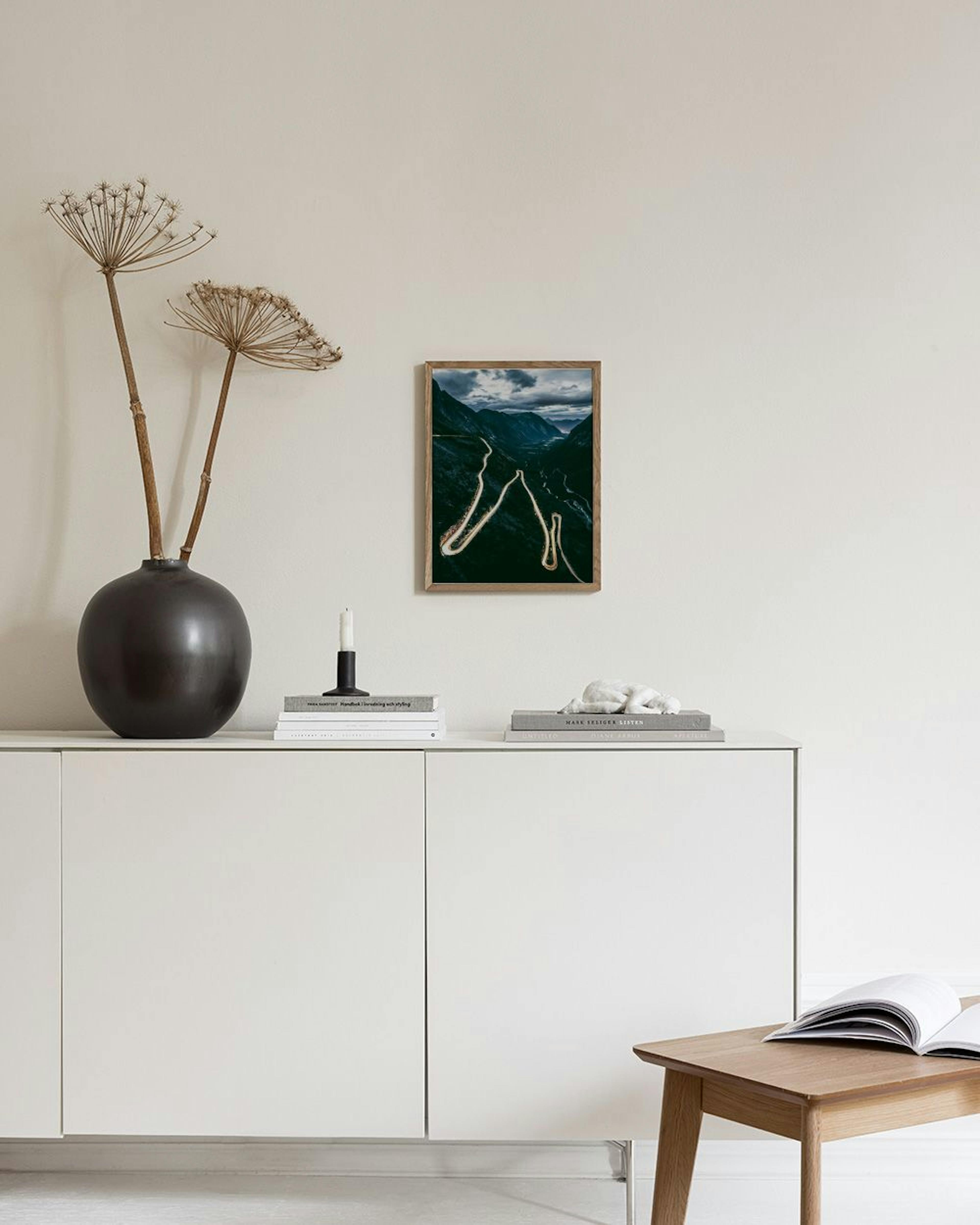 Poster van een kronkelende bergweg met lichtsporen bij schemering, hangend aan een donkere muur boven een salontafel.