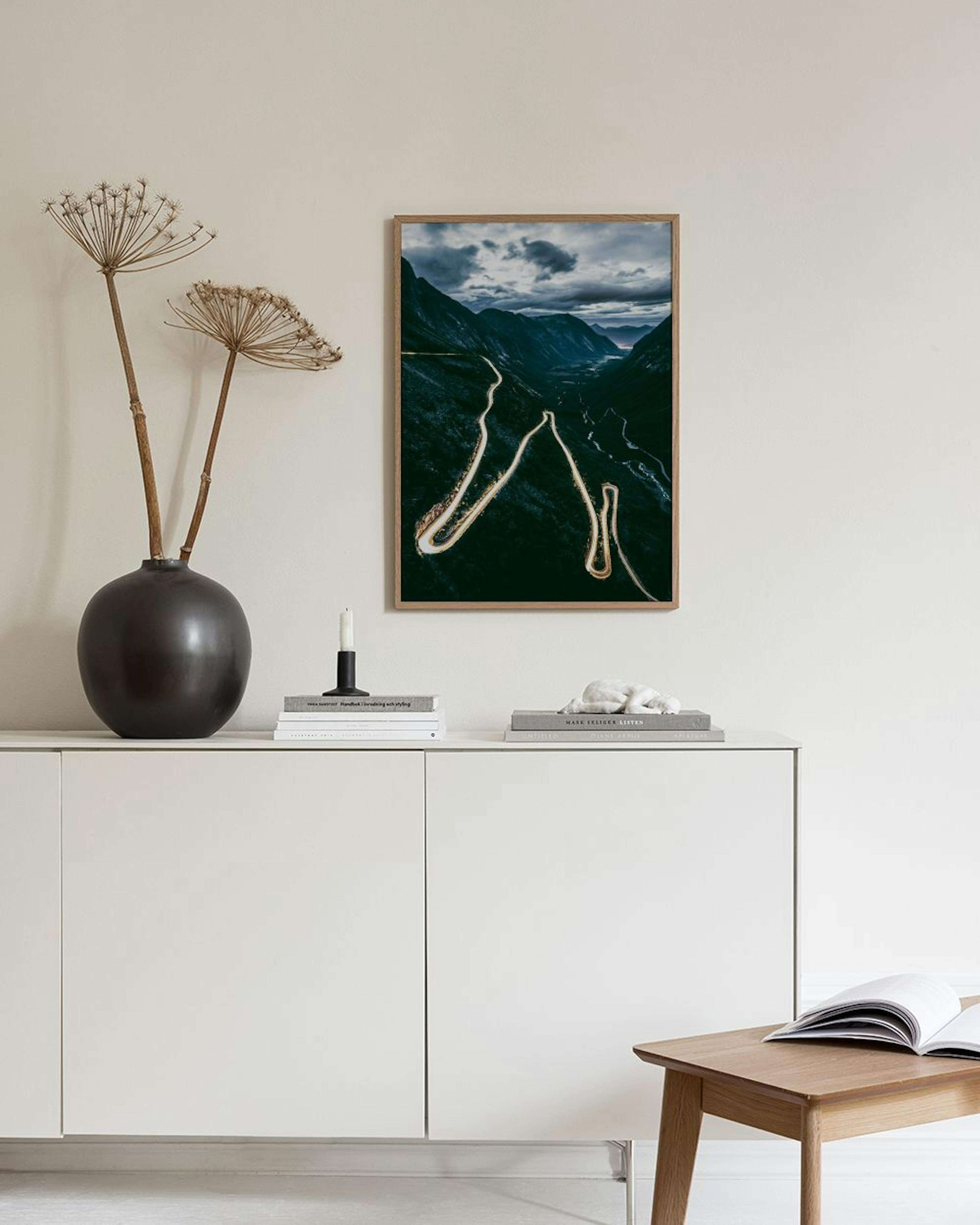 Póster de una carretera serpenteante en un valle montañoso bajo un cielo nublado, colgado en una habitación oscura con mesa y li
