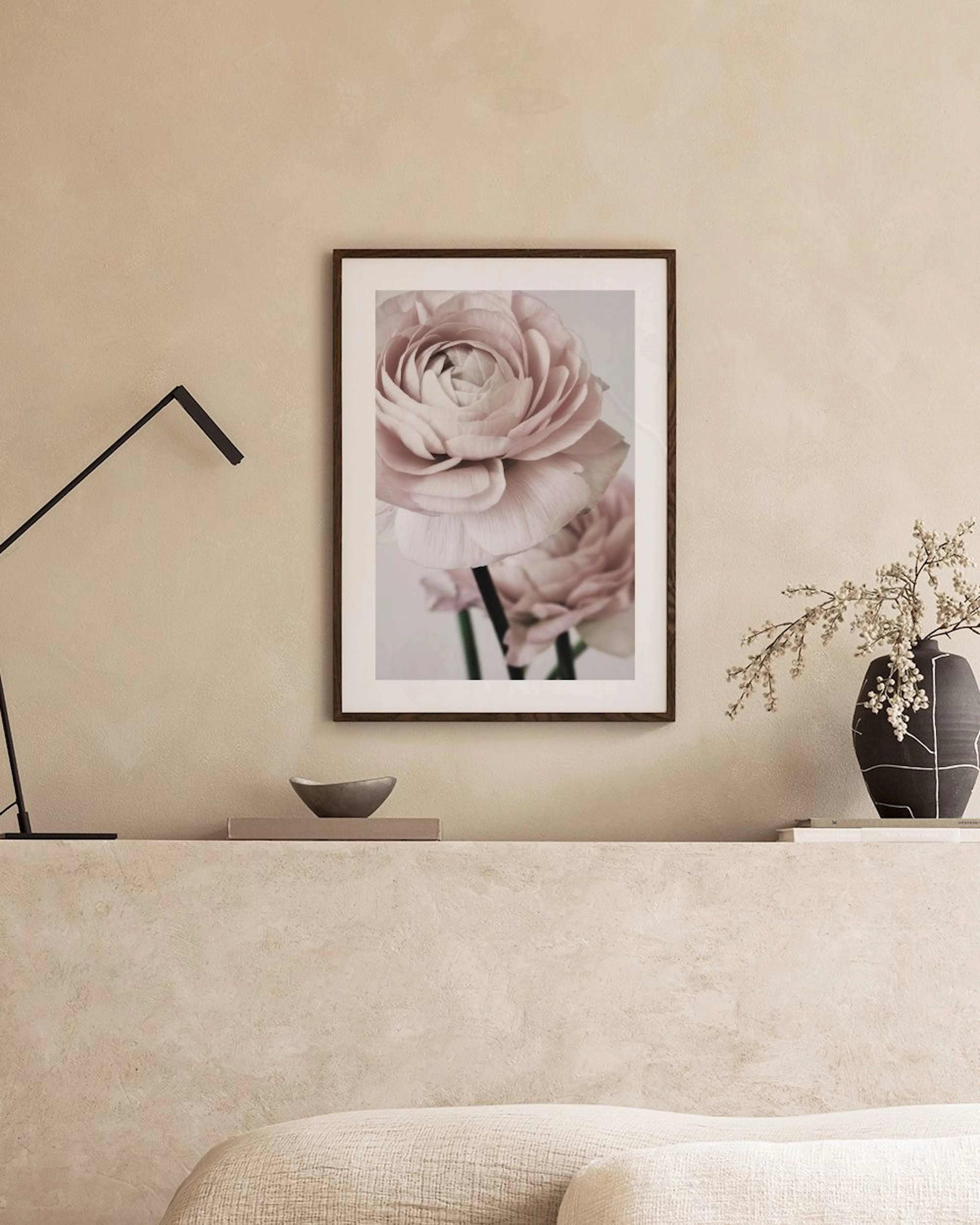 A print of delicate pink ranunculus flowers on a light background, hanging above a red cabinet with a beige vase and branch.