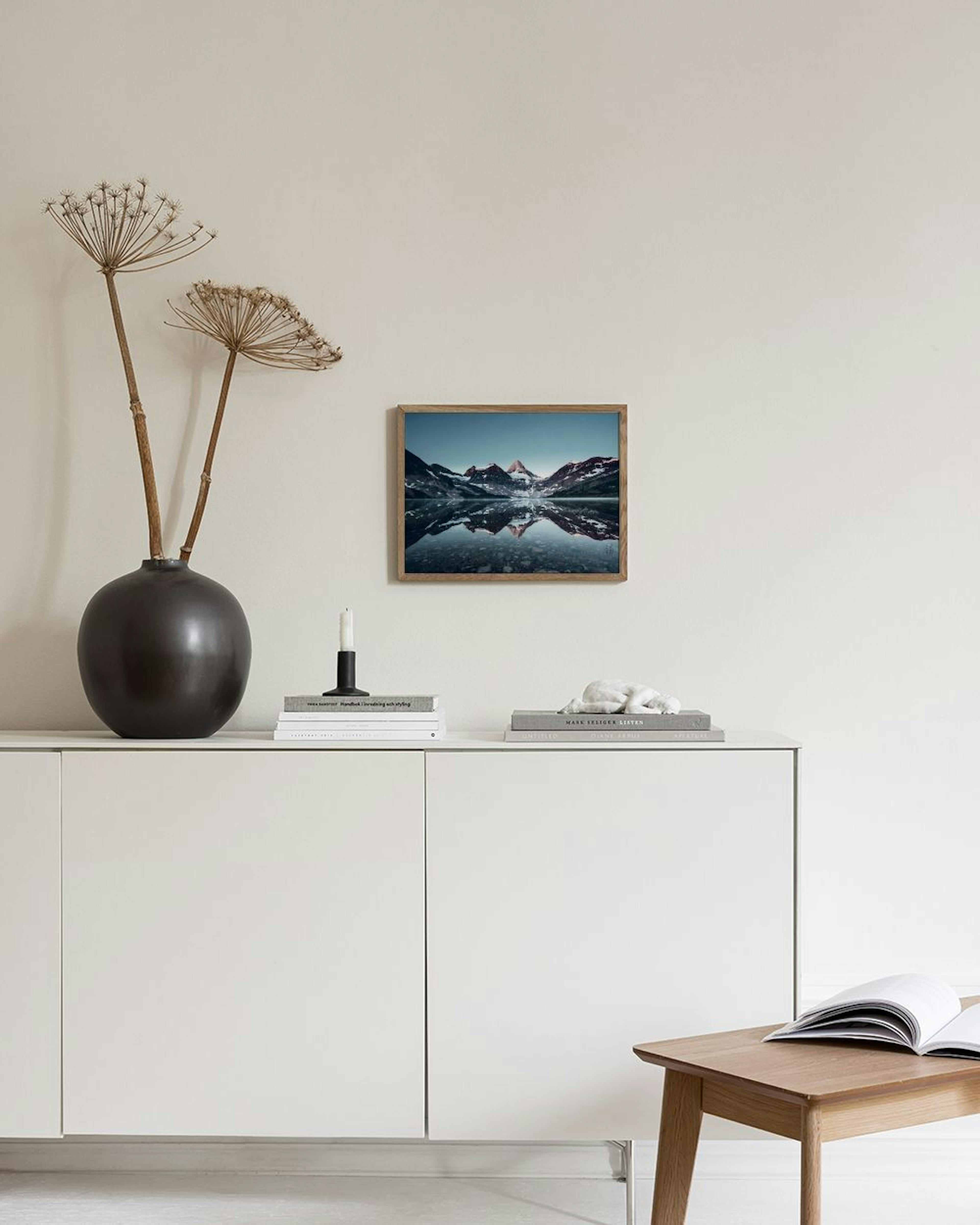 Poster mit Bergpanorama und Wasserspiegelung, über einem weißen Sideboard mit Kerze und Pflanze.