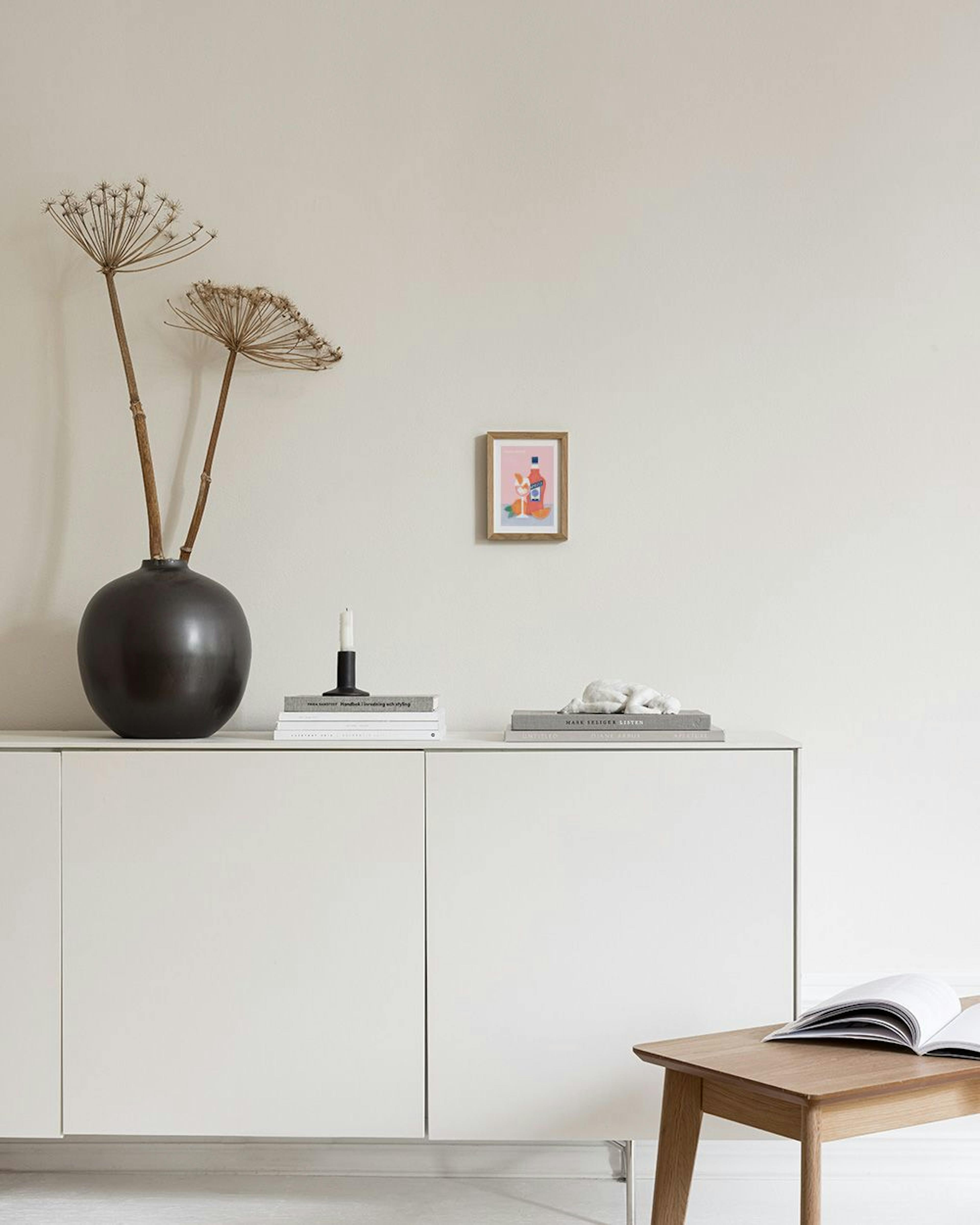 A colorful Spritz cocktail print with oranges and a bottle, displayed above a white side table with books and vases.