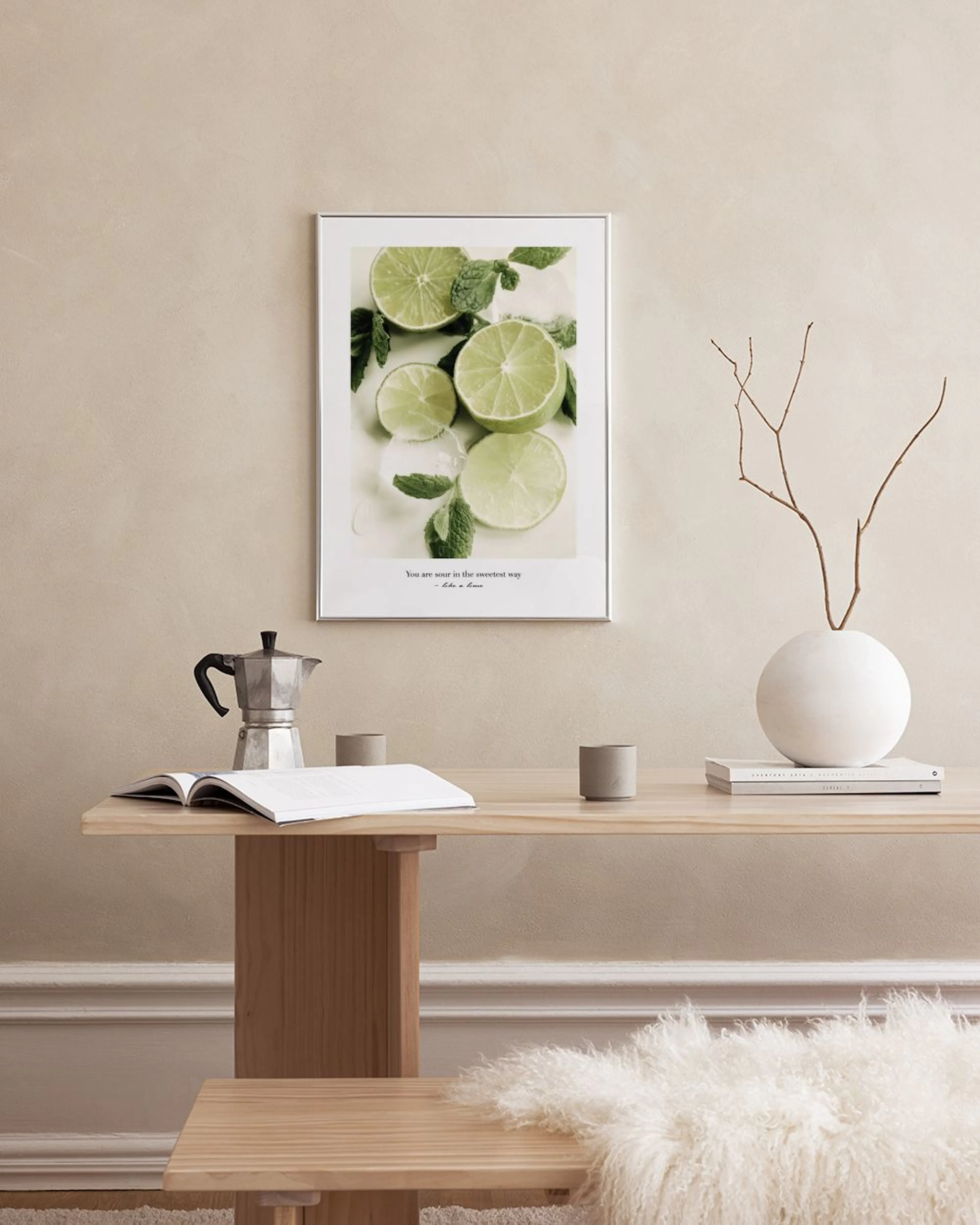 Póster con rodajas de lima, hojas de menta y hielo, colgado sobre un mueble blanco con tazas y botellas.