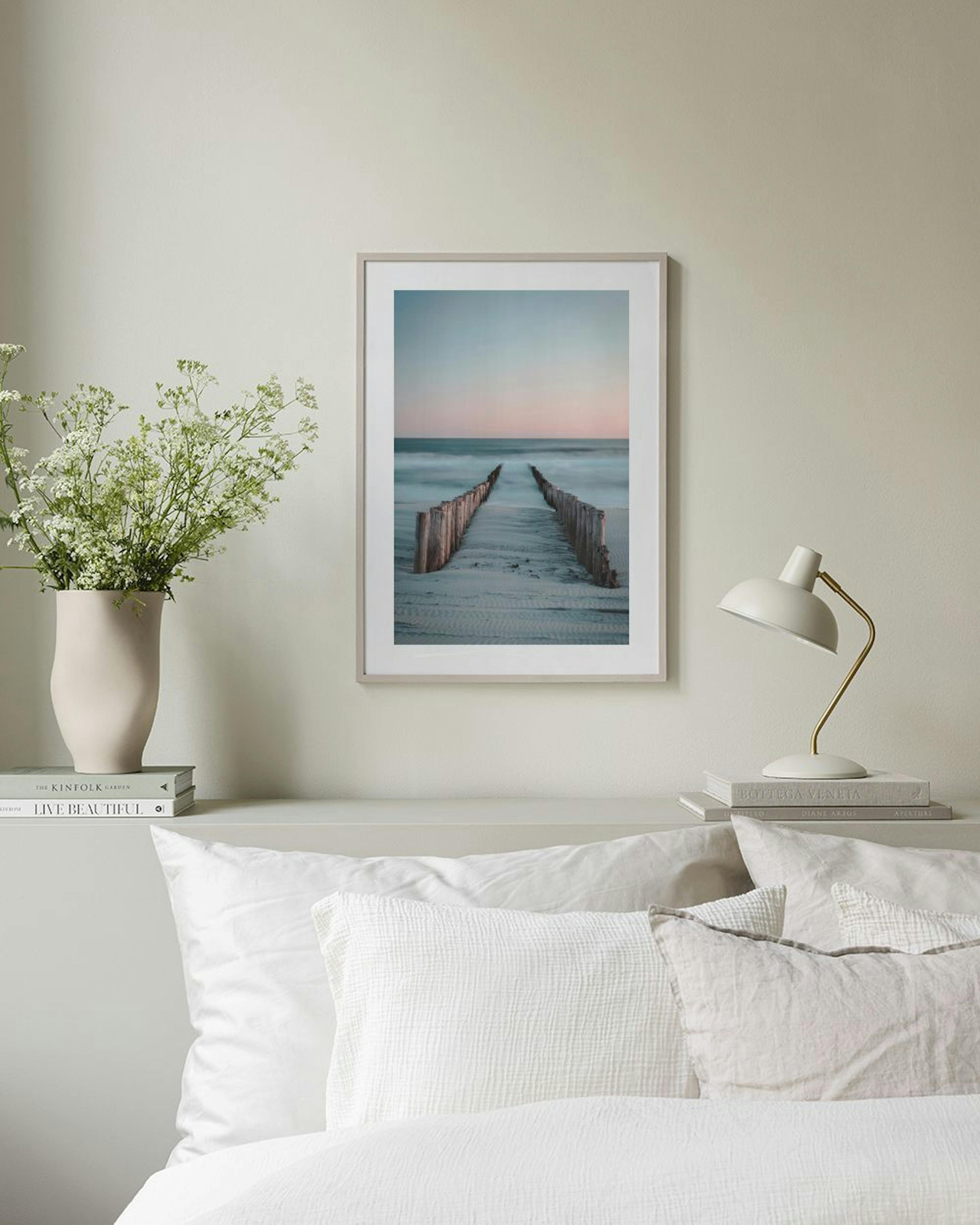Un póster de una pasarela de madera en la playa al atardecer, colgado en una pared gris junto a un sillón y una mesa auxiliar.