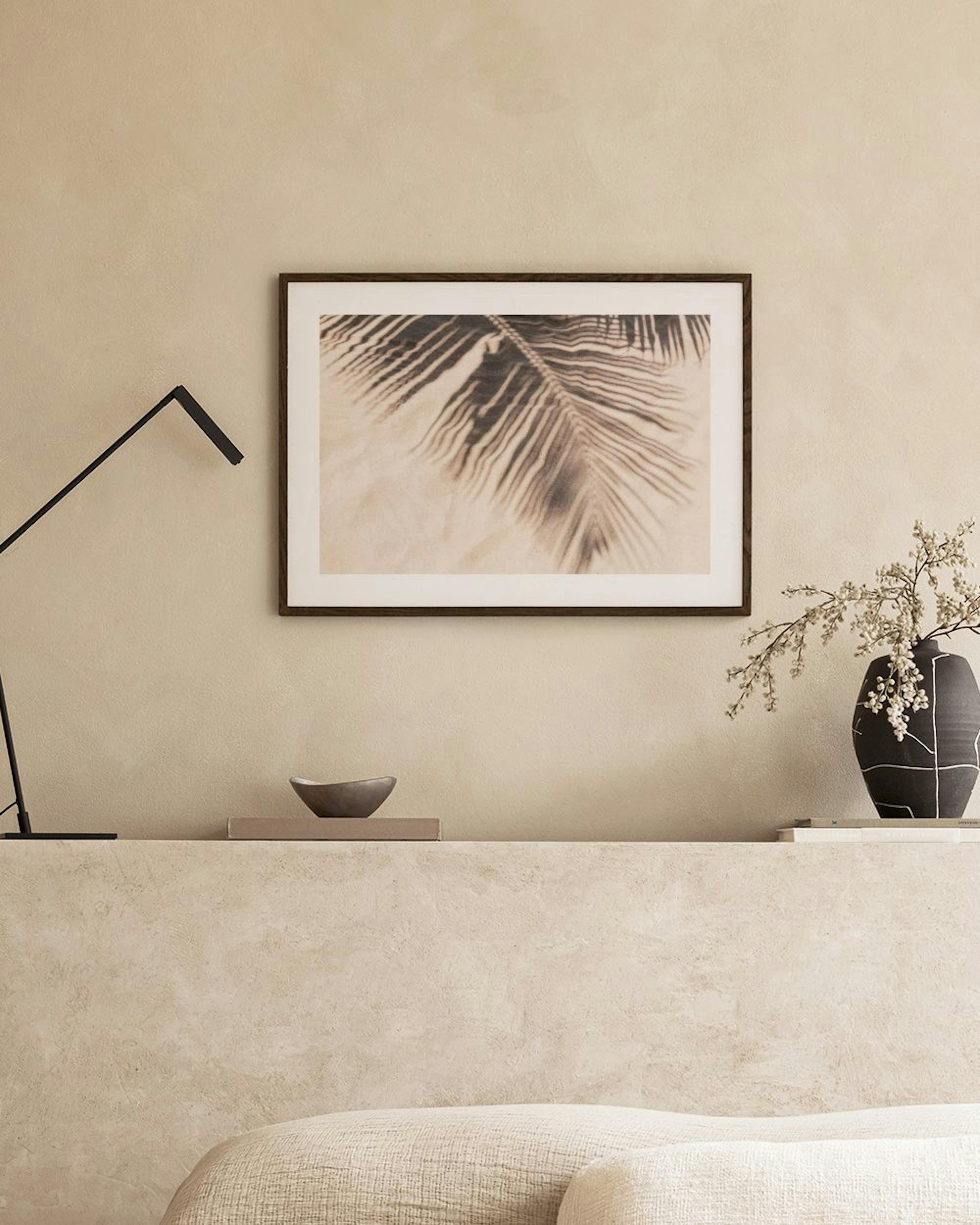 Sepia-toned print of palm fronds casting shadows on sand, displayed above a white cabinet with a vase and books.