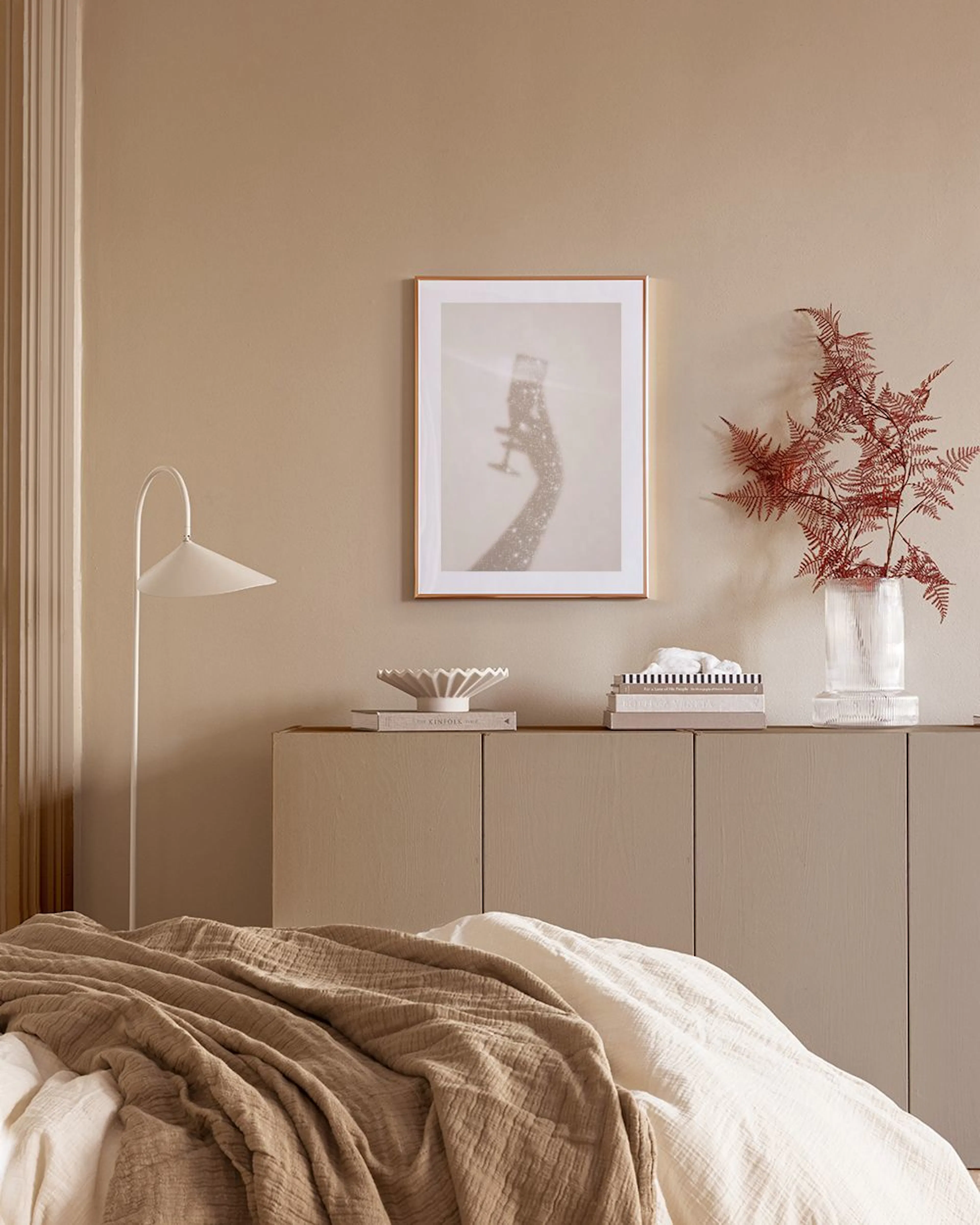 A print of a shimmering champagne glass and arm, displayed above a rattan cabinet with a sunburst mirror and vase.