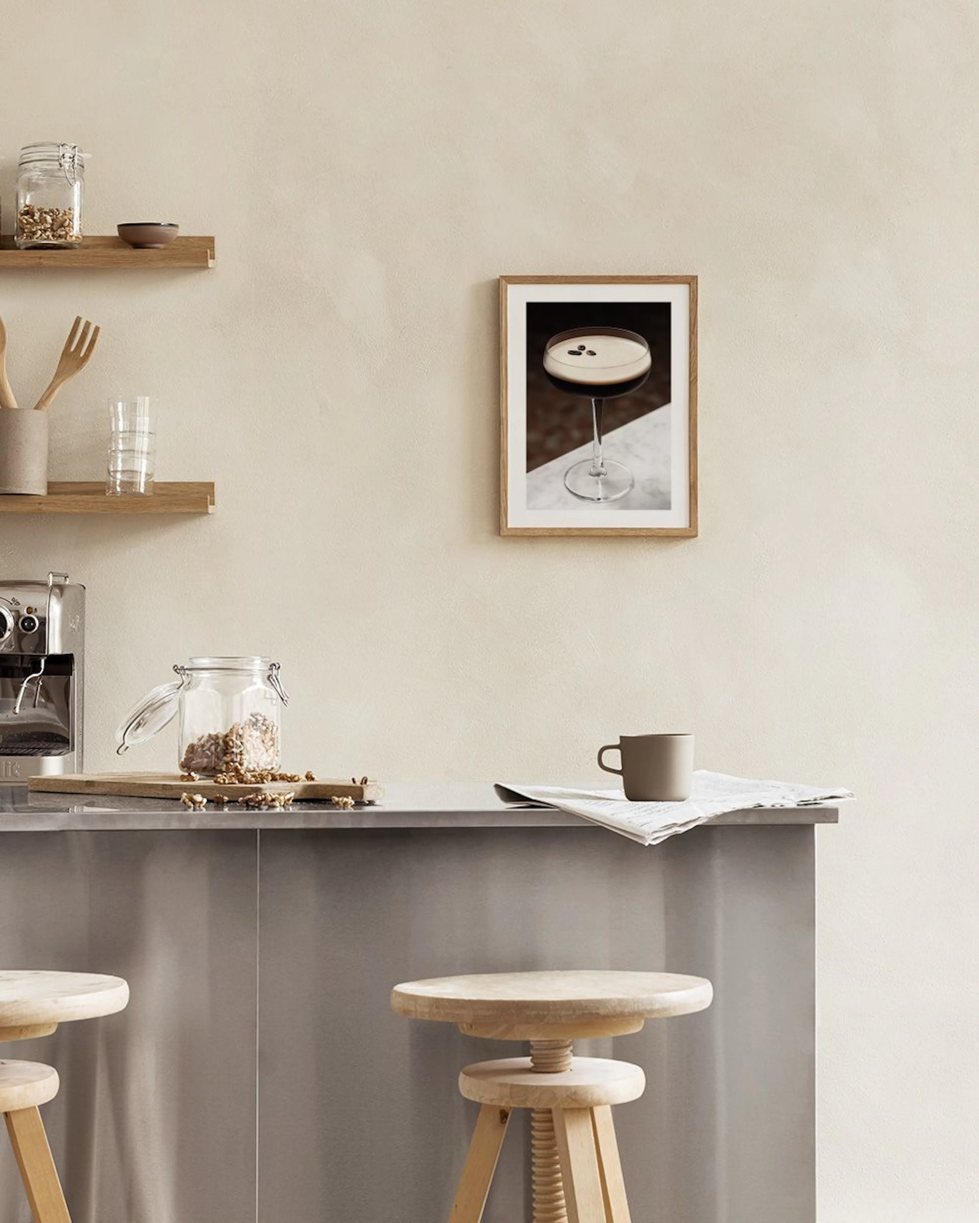 Póster de un espresso martini con granos de café, colgado sobre un mueble blanco con un jarrón y un taburete.