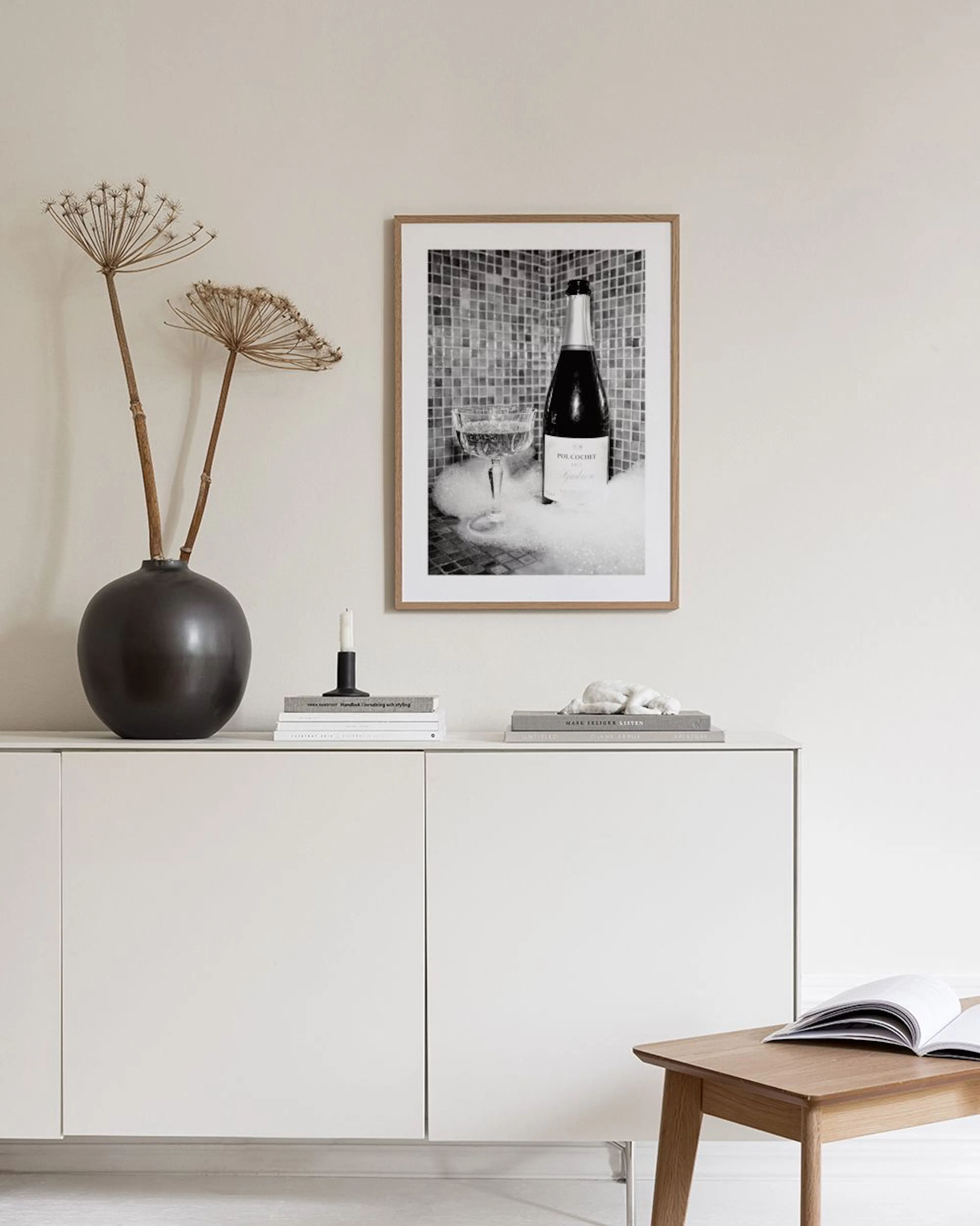 Zwart-wit poster van een champagnefles en glas in schuim, hangend boven een wit dressoir met vaas en krukje.