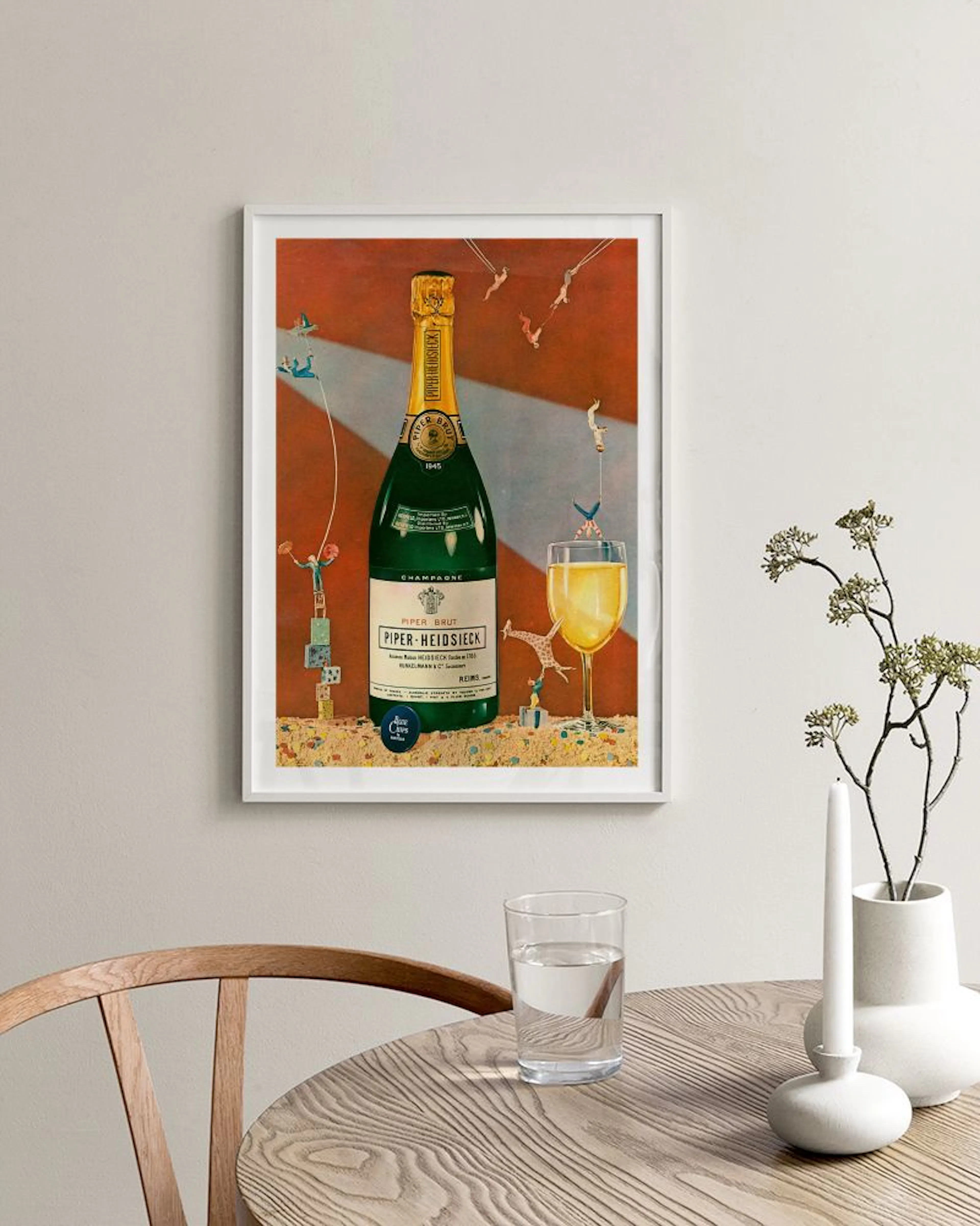 A vintage Champagne print with tiny acrobats on a bottle and glass, hanging above a chair and next to a black shelf.