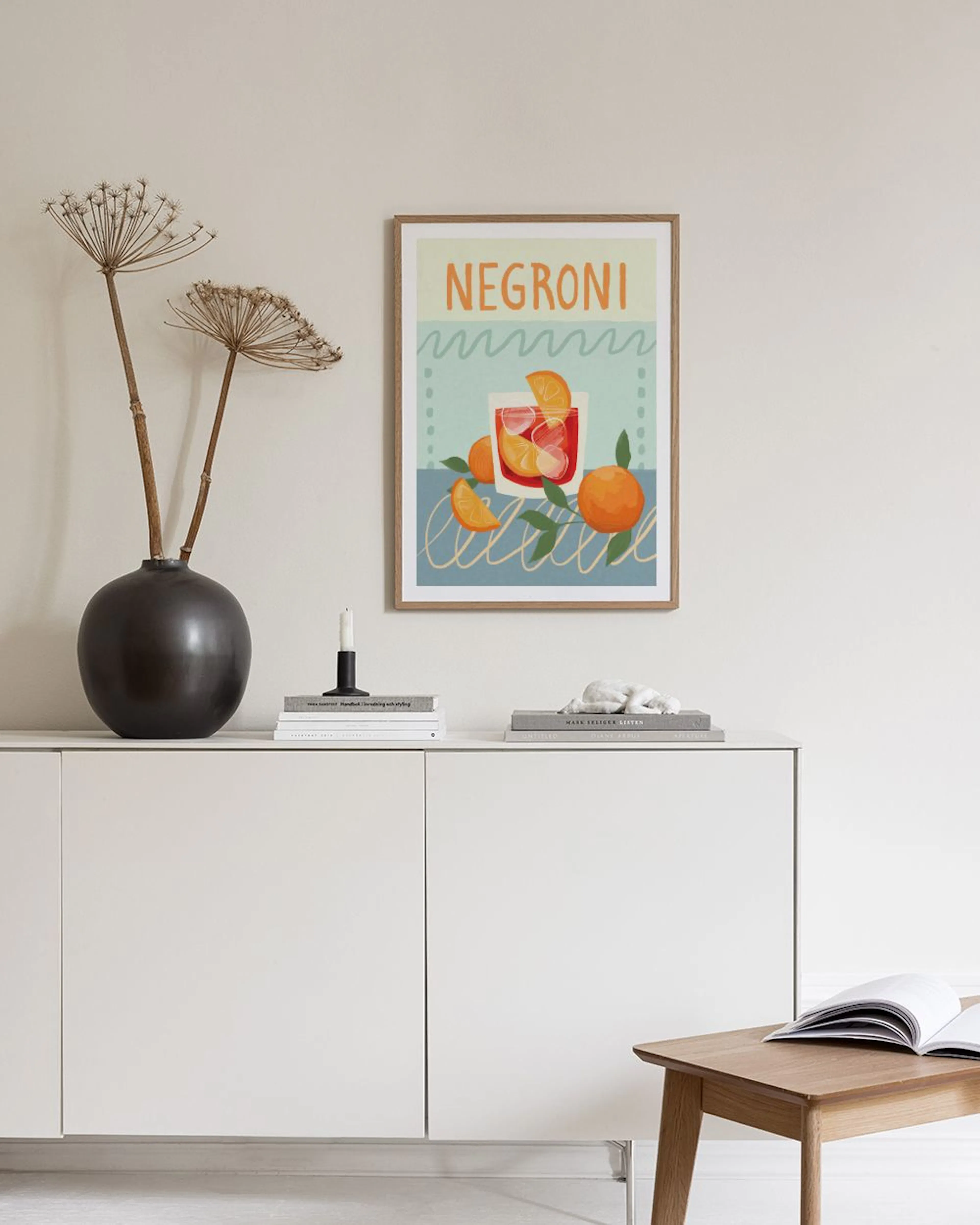 A Negroni drink print with oranges and green leaves, hanging above a black console table in a living room.