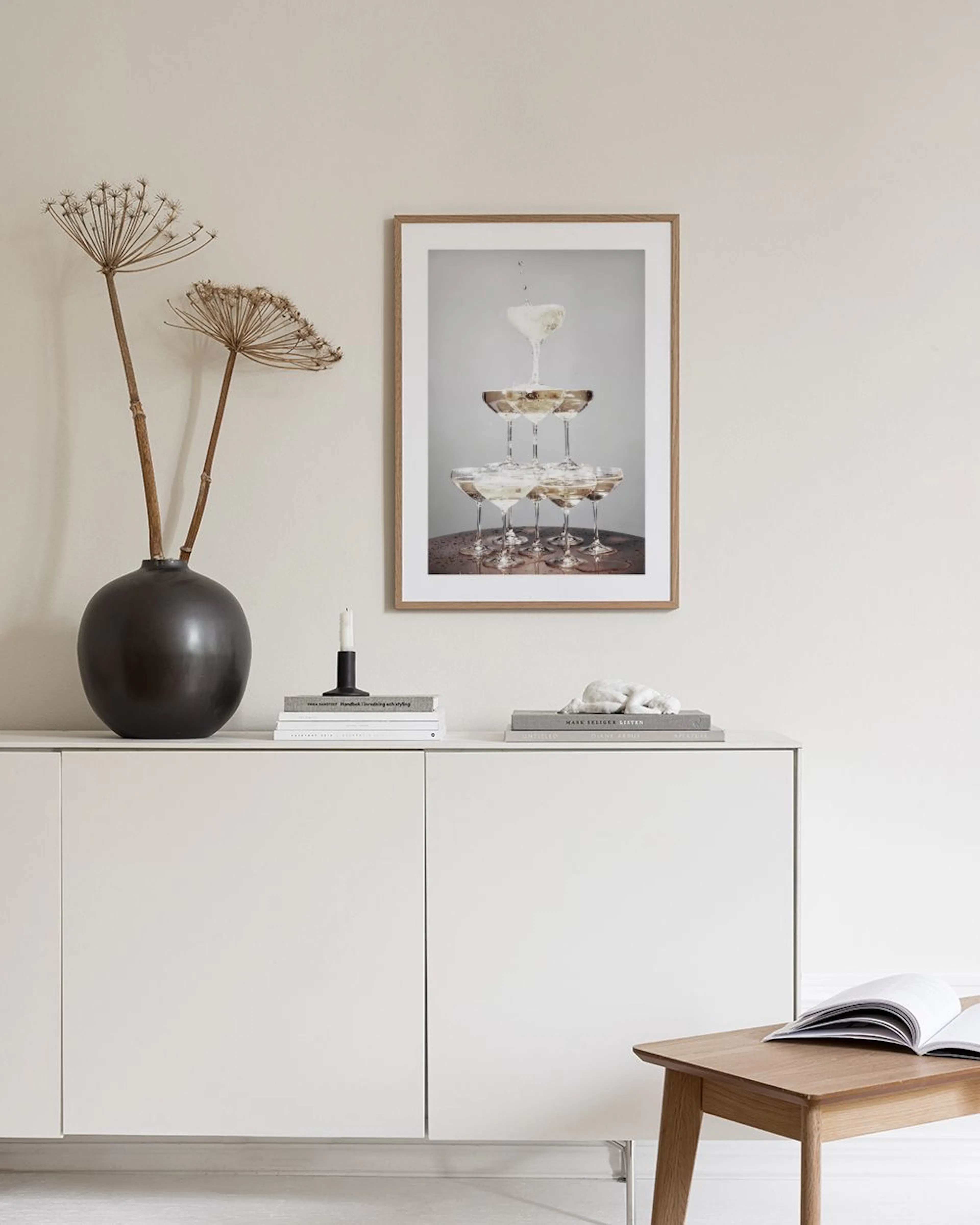 Poster mit Sektgläser-Pyramide, über einem goldenen Tablett mit Pflanzen und Kerzen, an einer dunkelblauen Wand.