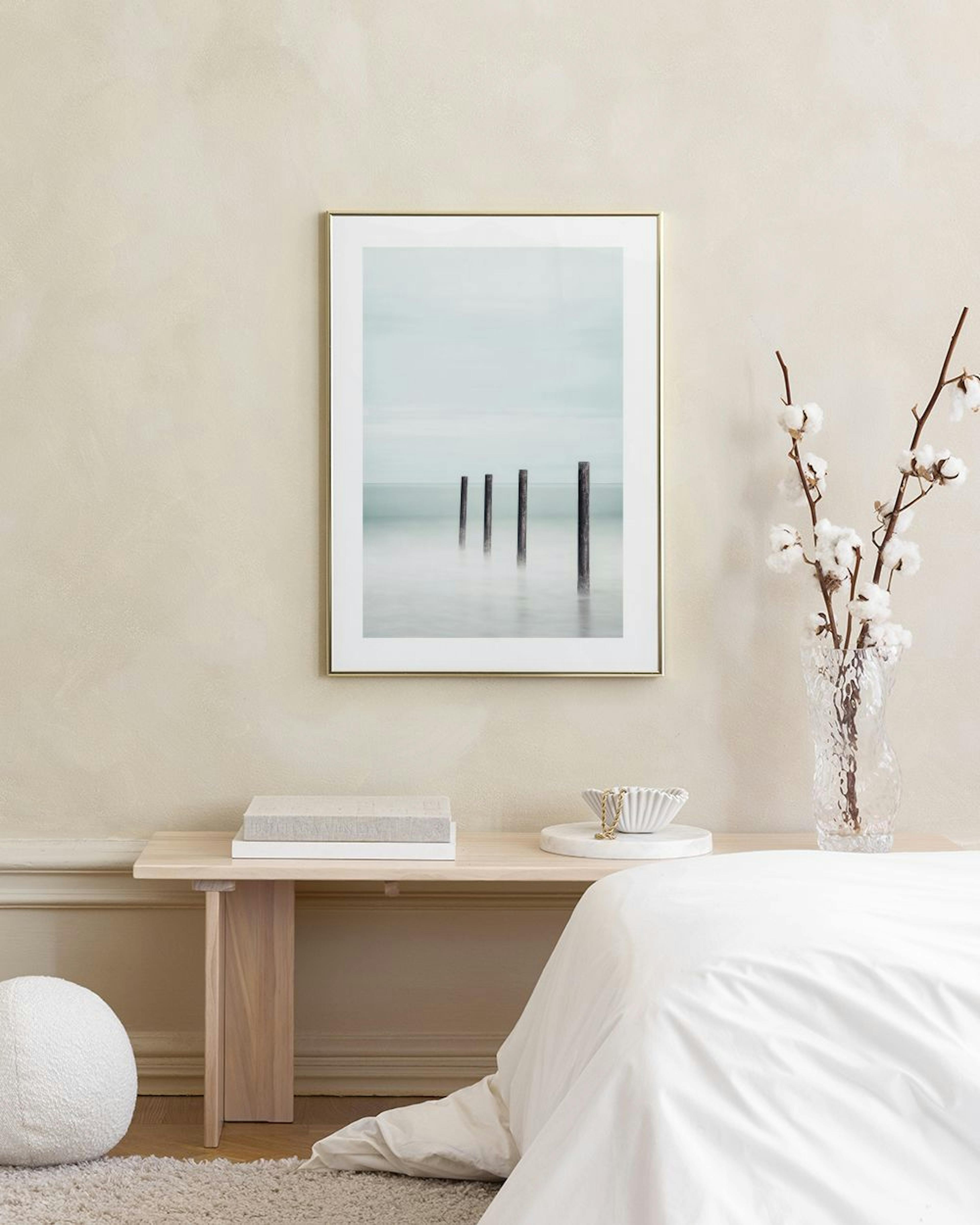 Poster mit vier Holzpfählen im nebligen Meer, hängt über einem Glastisch mit Büchern und Vase.
