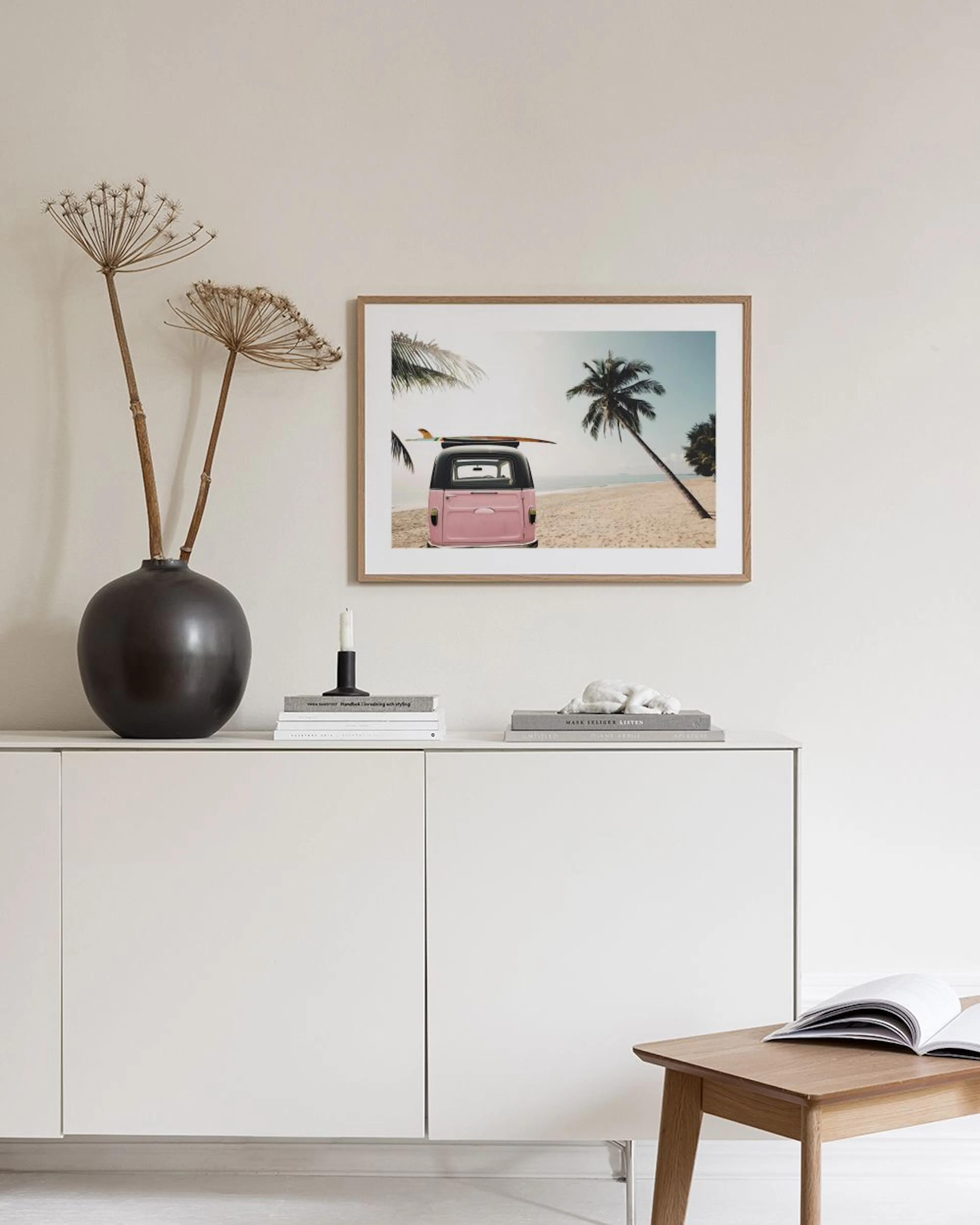 A print of a pink Kombi van with a surfboard on a tropical beach, hanging above a teal armchair and white pedestal.