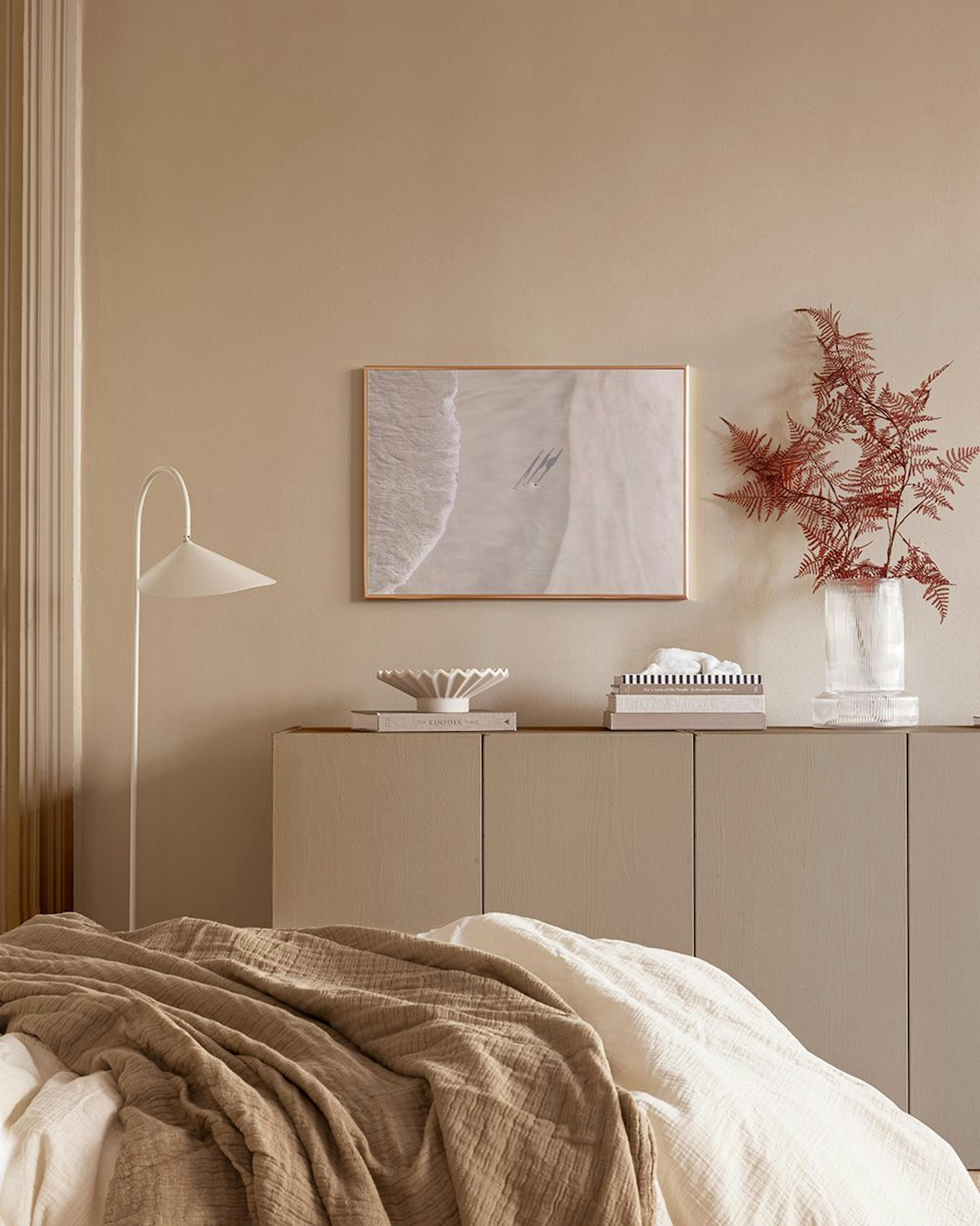 A print of a sandy beach with foamy waves and two people, displayed above a wooden cabinet with a lamp and vase.