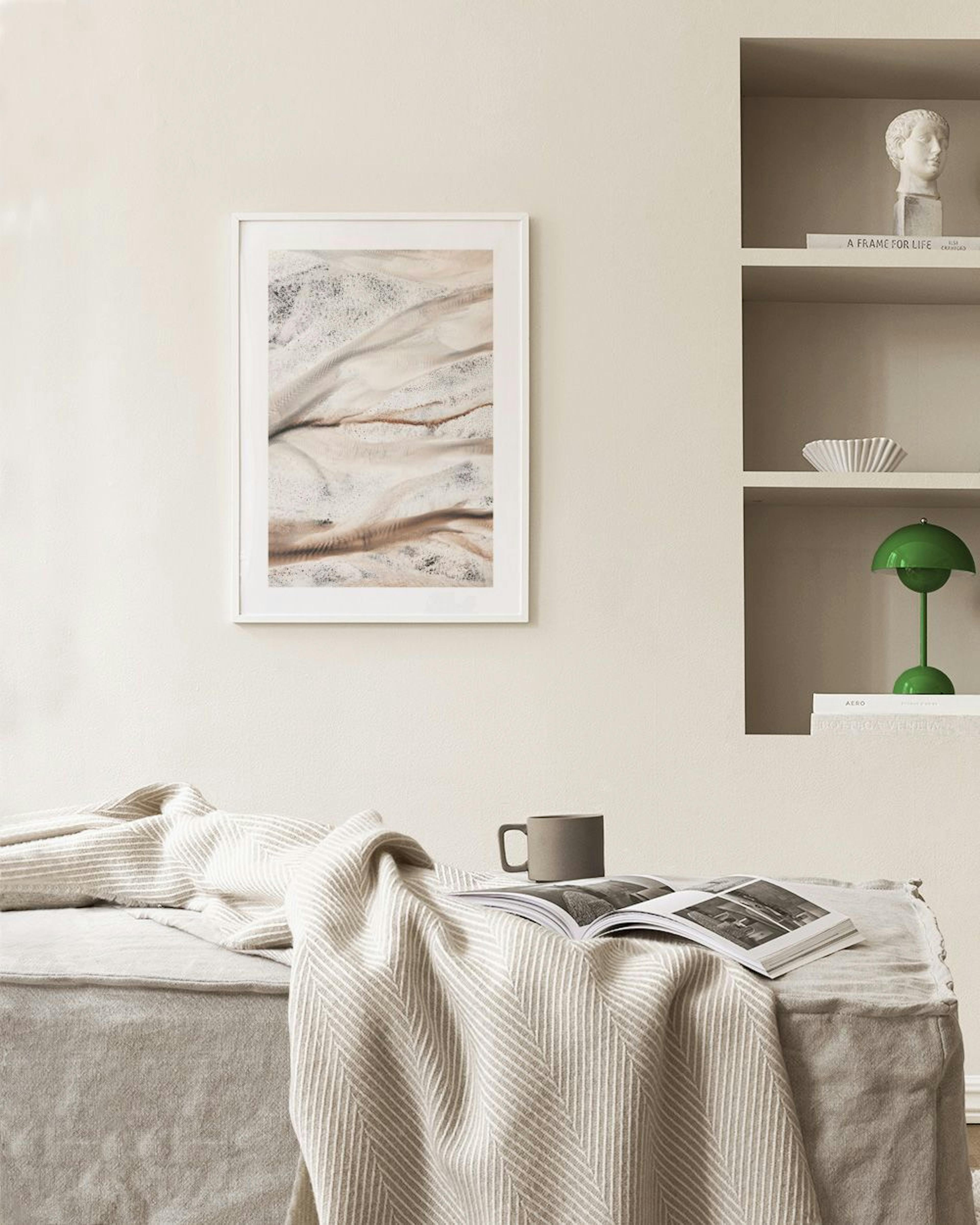 Póster de arte abstrato com texturas de areia e água em tons de bege e branco, pendurado sobre um banco de vime com decoração.