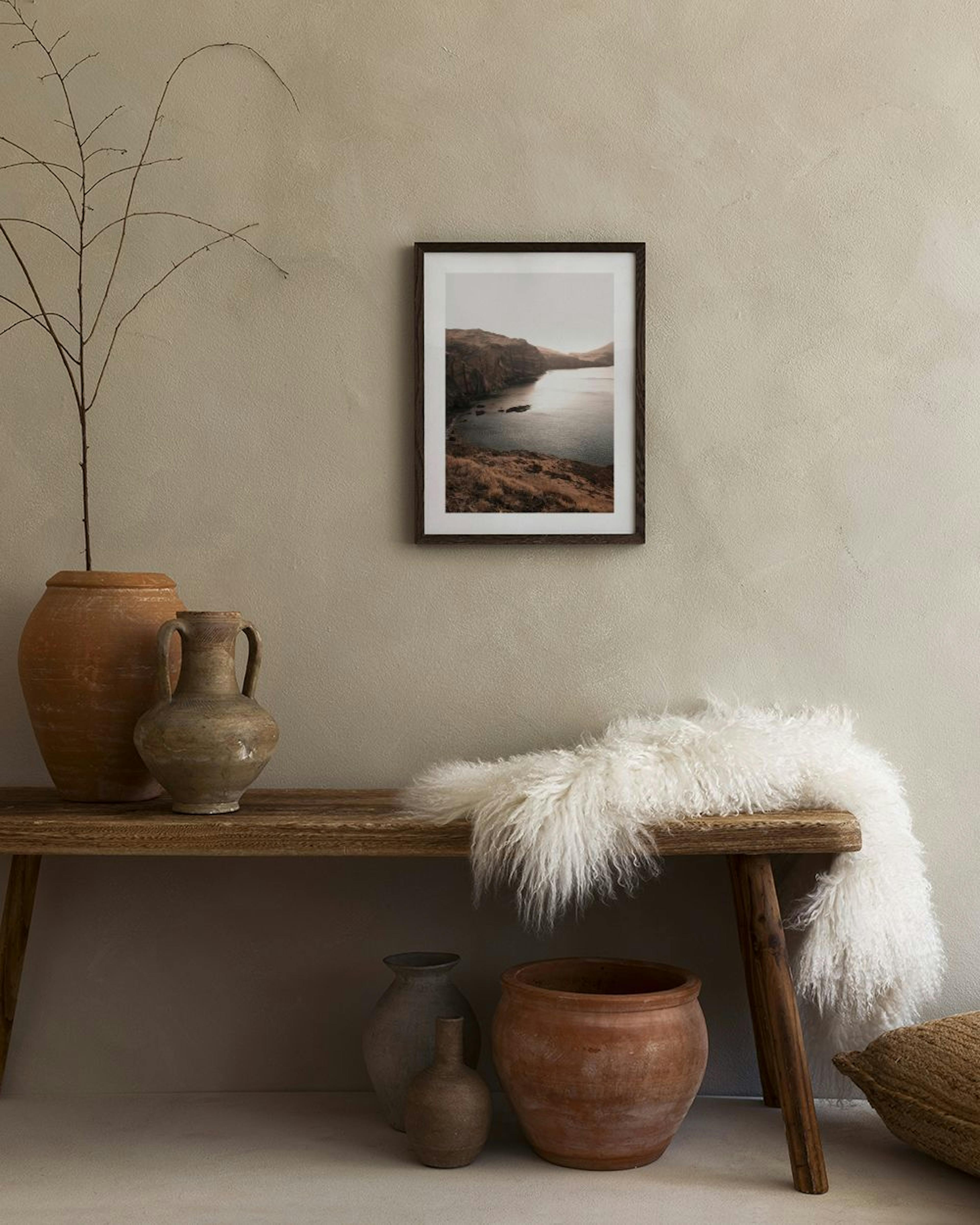 Poster van een ruige kustlijn met rotsen en een glinsterende zee, hangend naast een fauteuil met een vaas pampasgras.