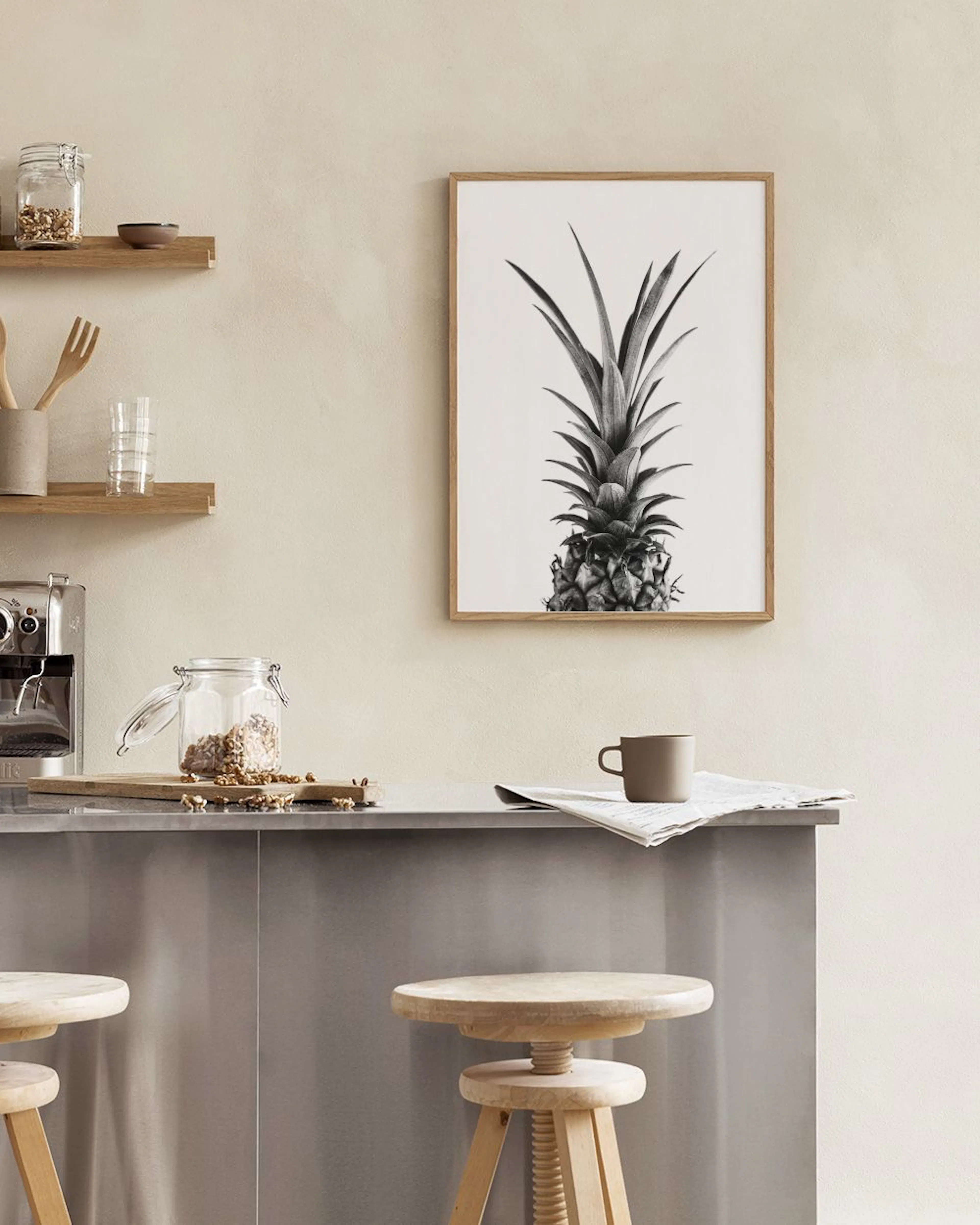 A black and white print of a pineapple hanging above a light wooden table with a book and coffee in a modern room.
