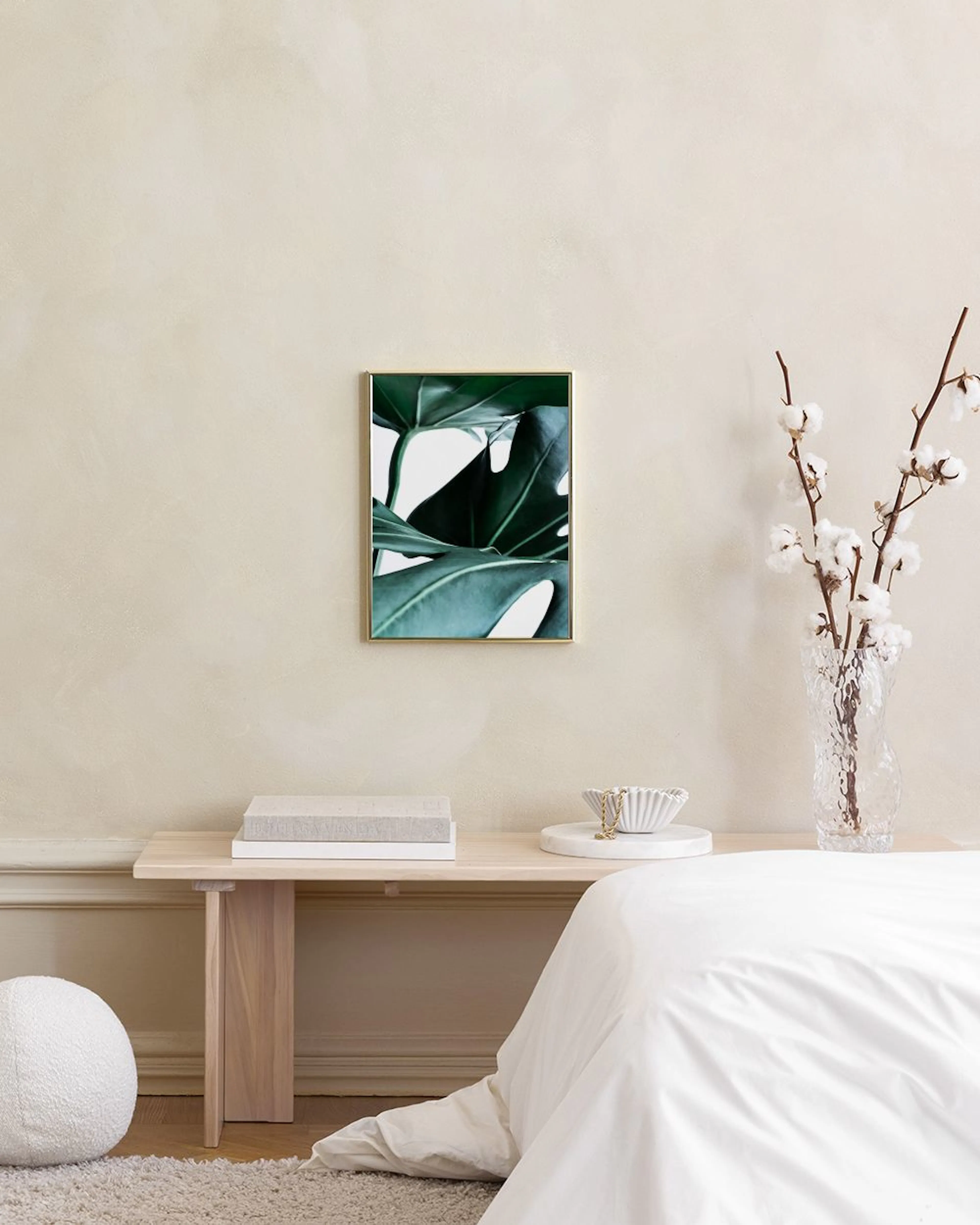 Poster mit Nahaufnahme von grünen Monstera-Blättern an einer weißen Wand über einem Hocker mit Büchern.