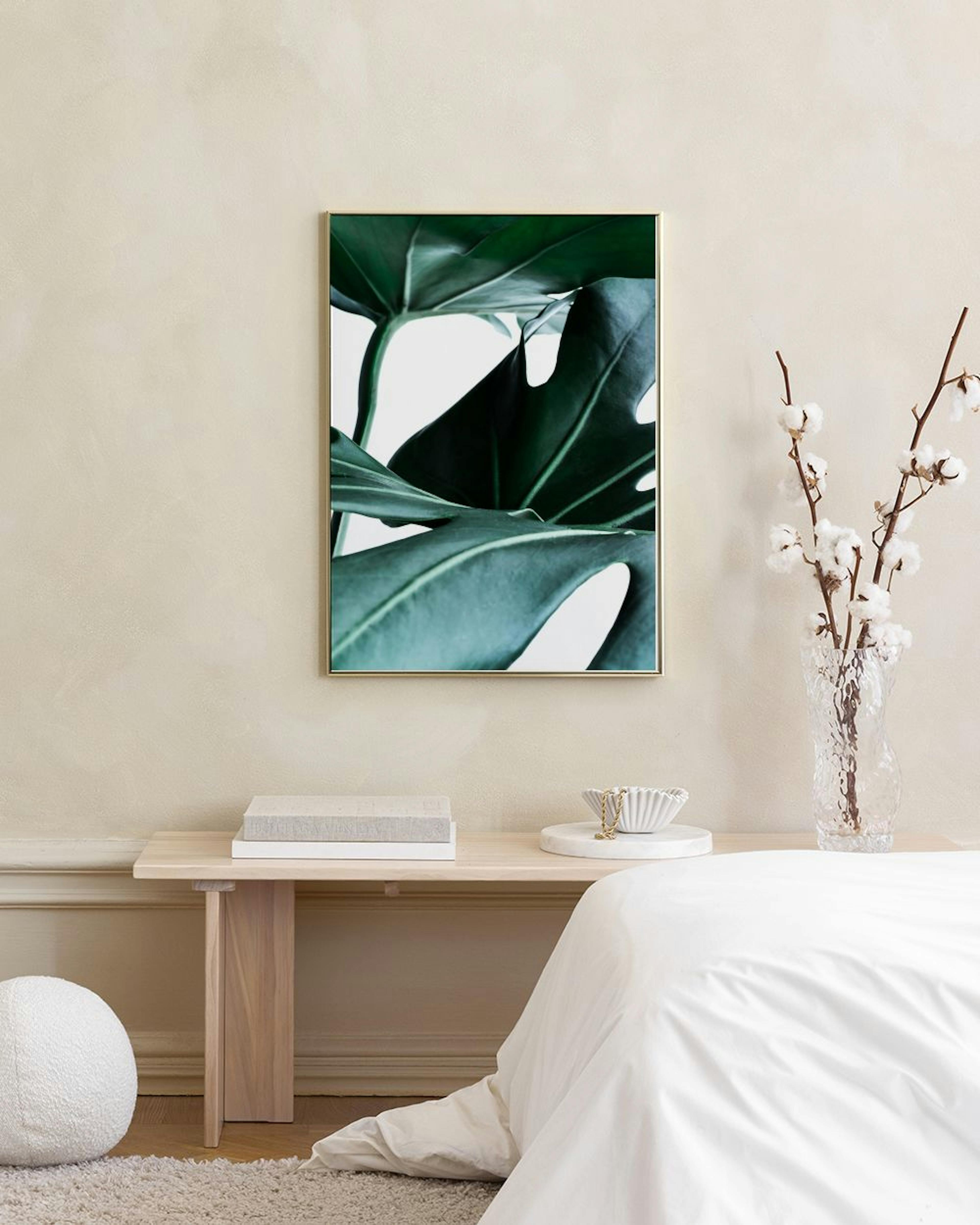 Poster con foglie di Monstera verde scuro e chiaro, appeso sopra uno sgabello con libri su una parete bianca.