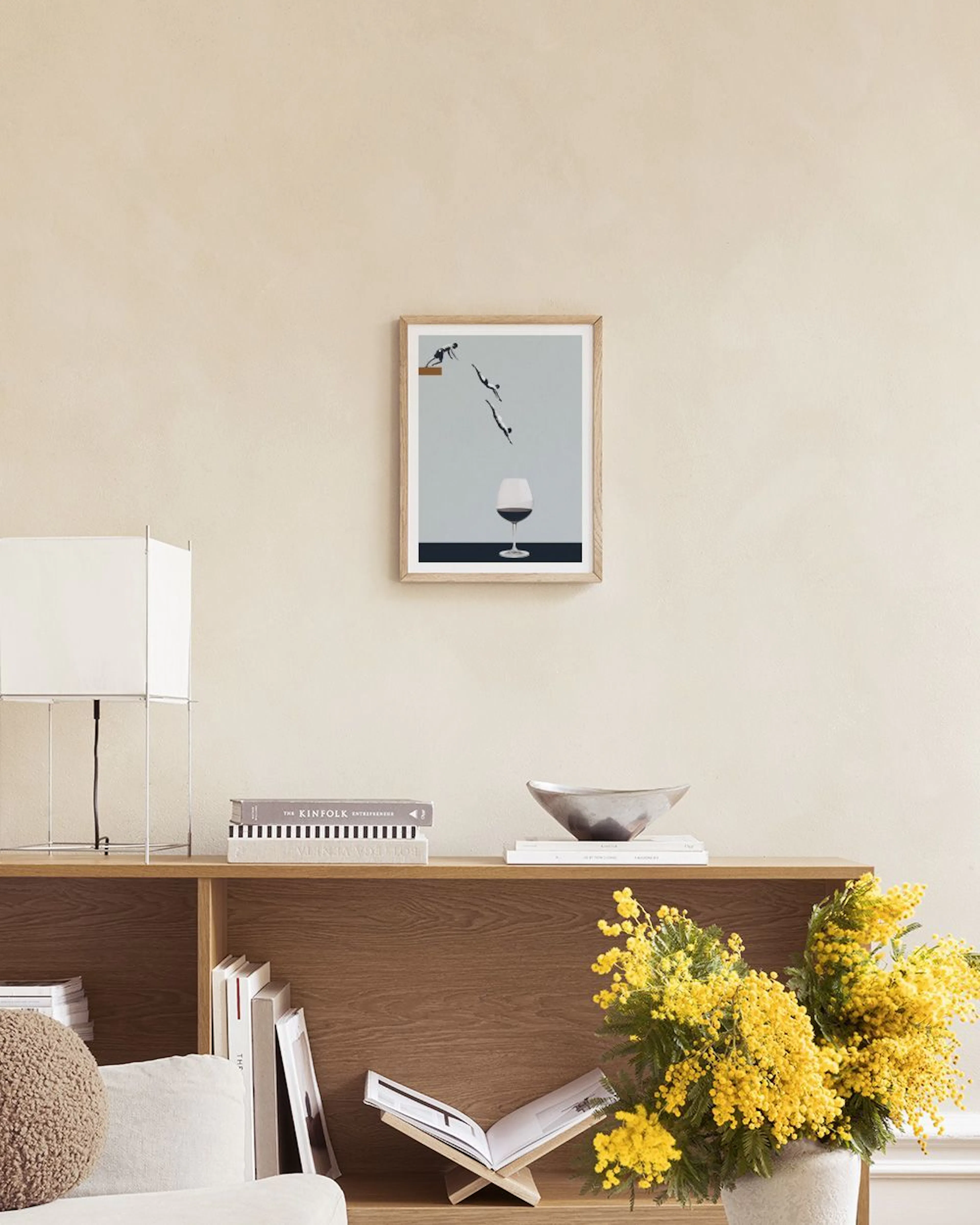 A print of people diving into a wine glass, hanging on a white wall above a cork stool with books.