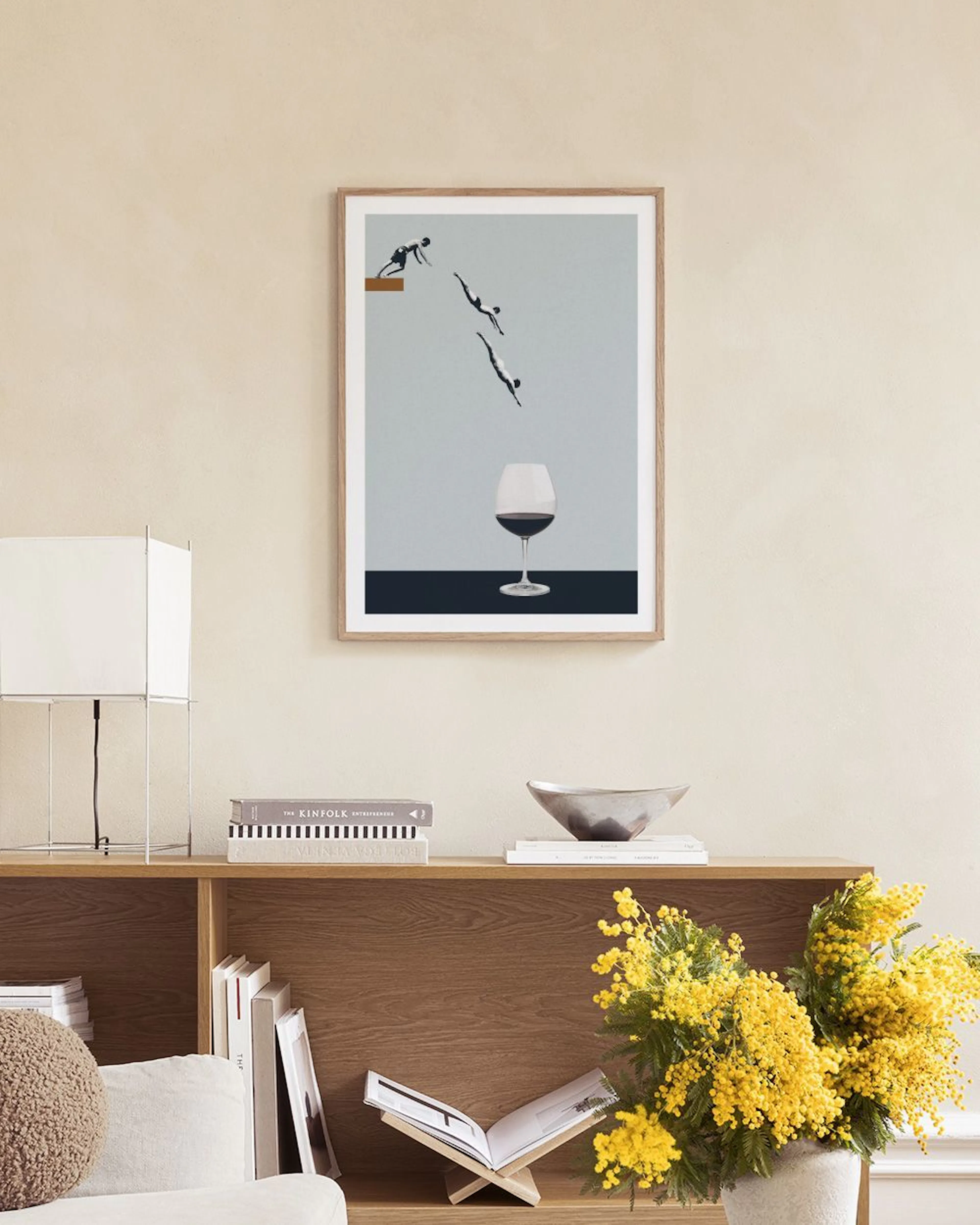 Póster de buceadores saltando hacia una copa de vino, colgado sobre un taburete con libros en una habitación moderna.