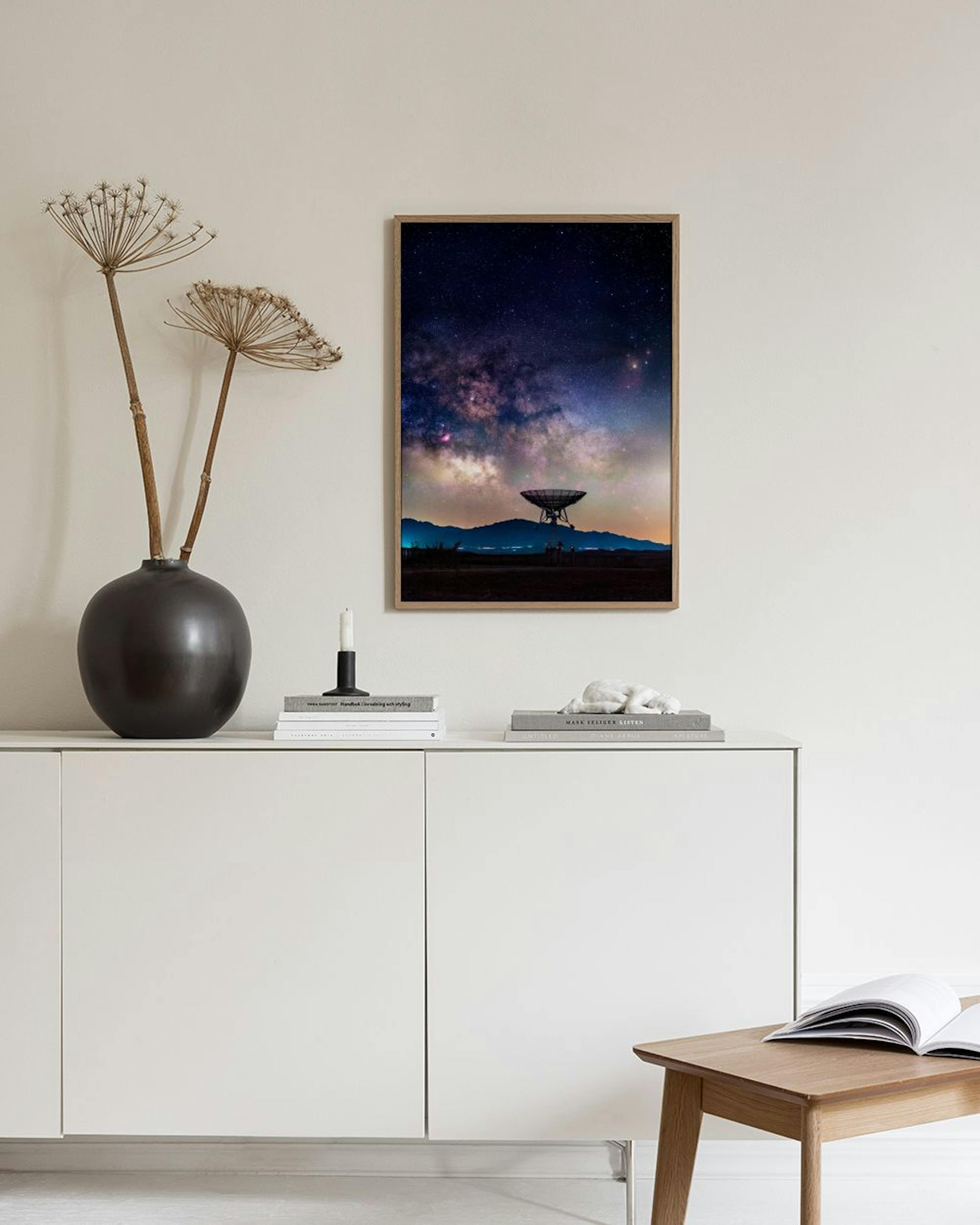 Poster con radiotelescopio su una collina illuminata dalla Via Lattea e stelle, appeso sopra una credenza rosa con vasi e libri.